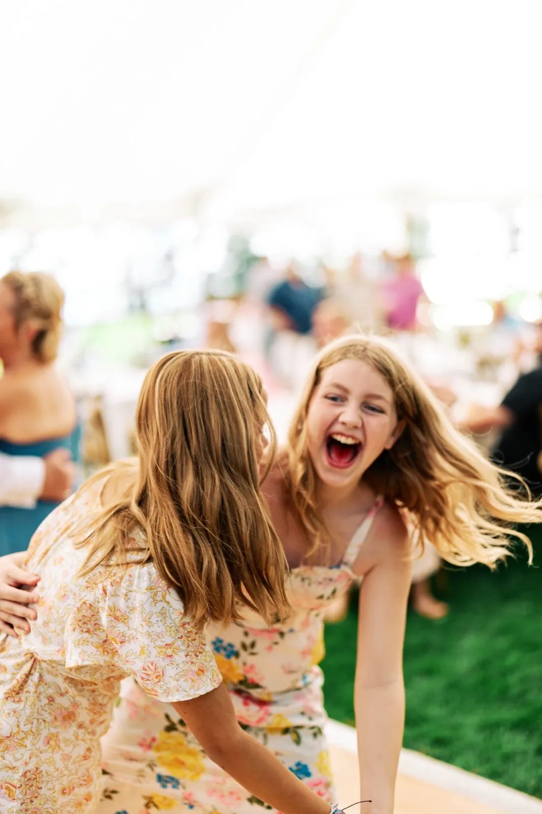 A young guest with windblown hair laughs open-mouthed while a second guest in a floral dress leans toward her under a reception tent. — Tim Larsen Photography, Brainerd Lakes MN