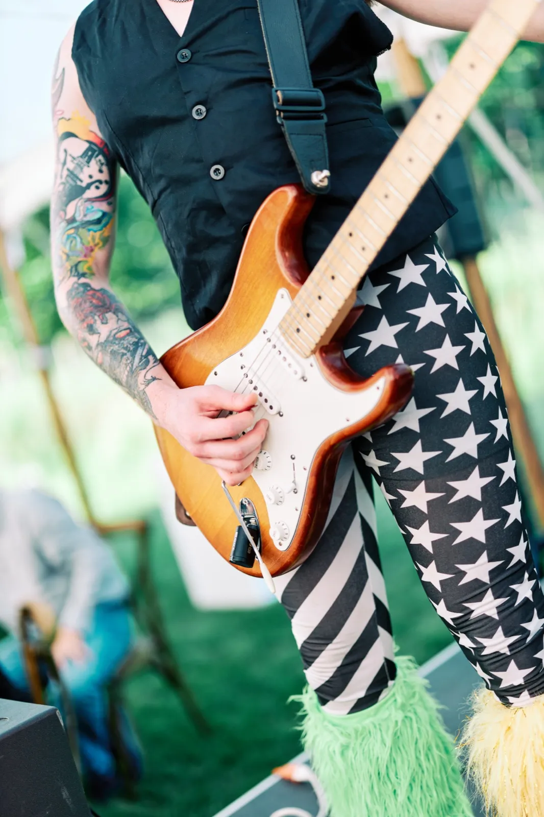 A tattooed guitarist in a black vest and star-patterned leggings with green and yellow feathered boots plays a sunburst electric guitar at an outdoor reception. — Tim Larsen Photography, Brainerd Lakes MN