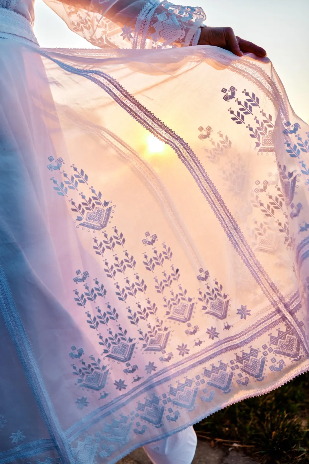 The bride holds an embroidered sheer veil up against the setting sun, the fabric's lace patterns illuminated as light passes through. — Tim Larsen Photography, Minnesota