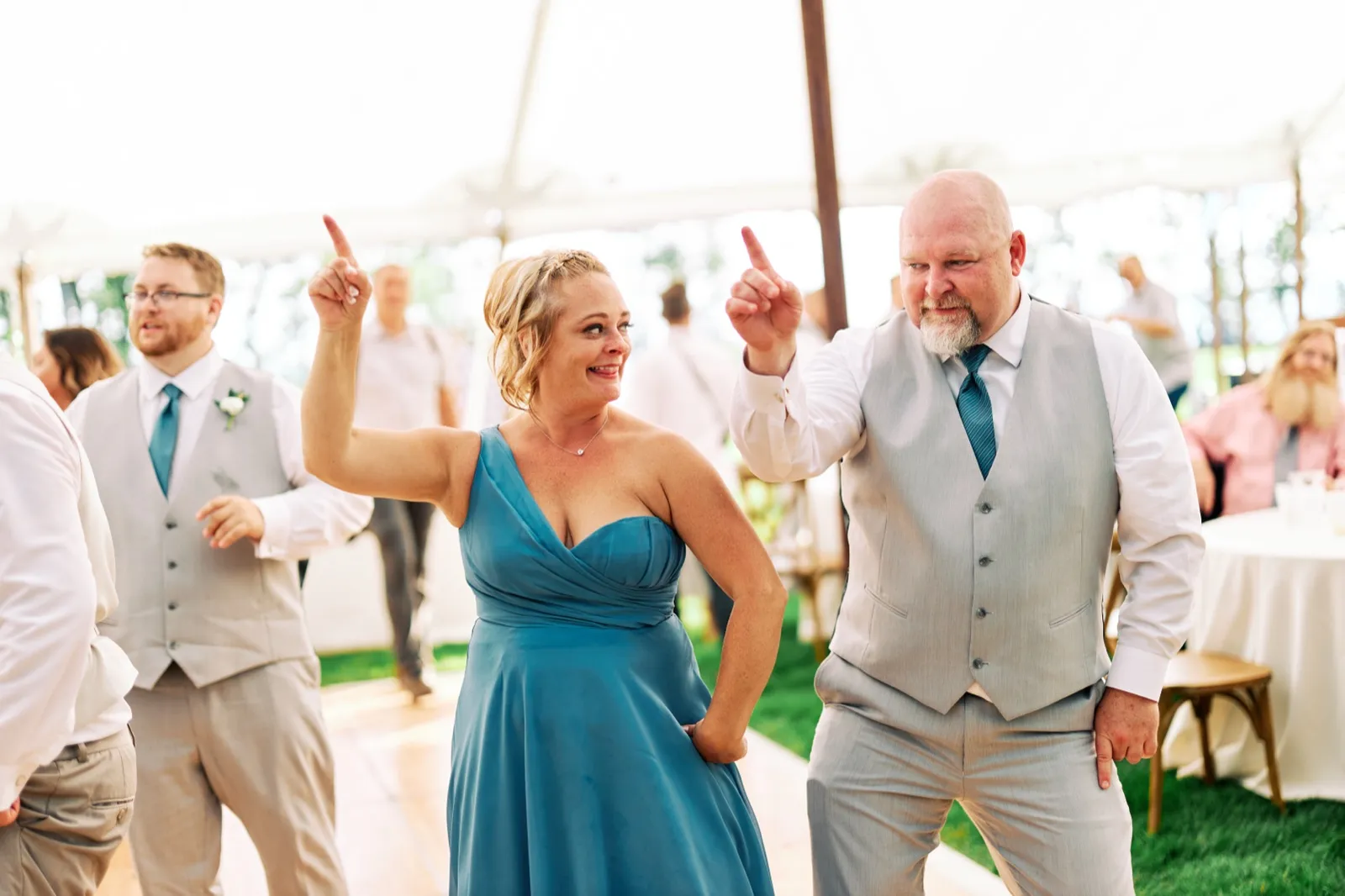A bridesmaid in a blue dress and a groomsman in a gray vest both point upward in sync as they dance under a reception tent with guests visible behind them. — Tim Larsen Photography, Brainerd Lakes MN