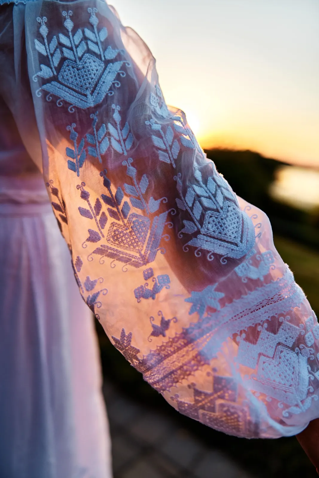 A close-up of the bride's sheer embroidered sleeve glowing in golden backlight with a lake and sunset visible in the soft background. — Tim Larsen Photography, Minnesota