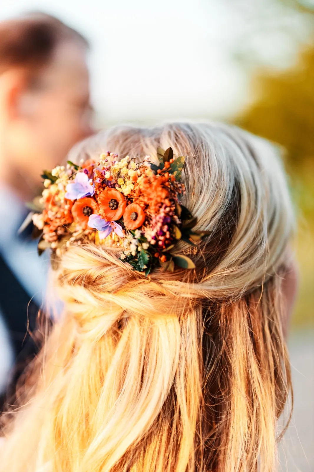 A close-up from behind shows the bride's braided hair adorned with a cluster of orange poppies, purple blooms, and dried florals, with the groom softly out of focus in the foreground. — Tim Larsen Photography, Minnesota