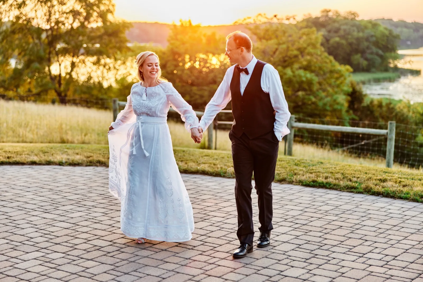 The bride and groom walk hand in hand across a stone patio at sunset, with a lake and treeline glowing behind them. — Tim Larsen Photography, Minnesota