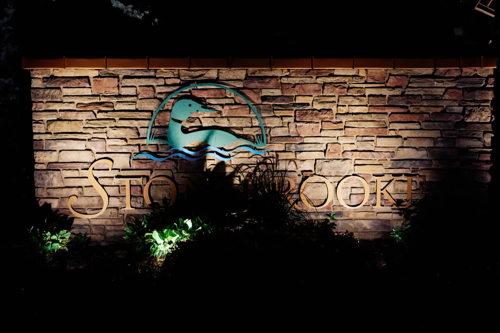 The illuminated Stonebrooke stone entrance sign featuring a loon emblem stands lit against a black night sky. — Tim Larsen Photography, Twin Cities MN