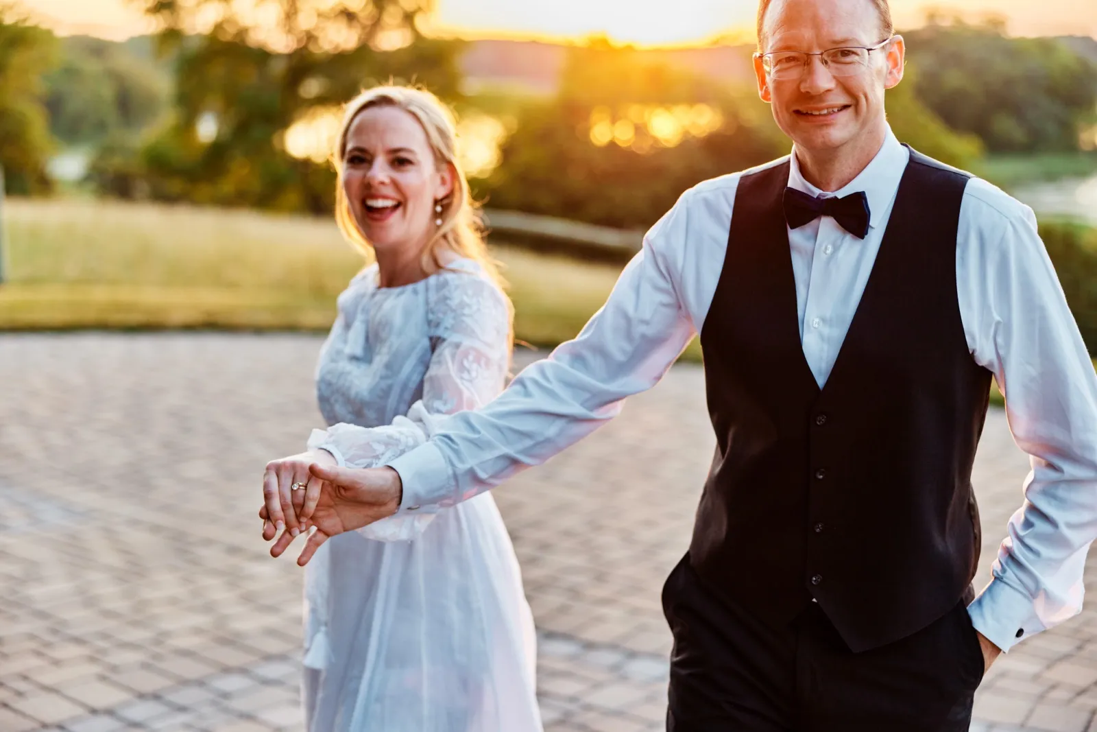 The bride laughs as she walks hand-in-hand with the groom across a stone patio, the setting summer sun glowing behind them. — Tim Larsen Photography, Minnesota