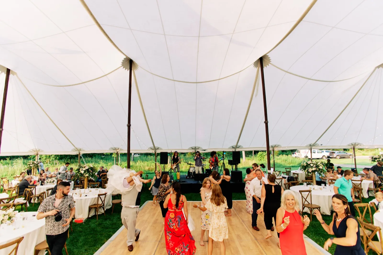 An ultra-wide view inside a large white sailcloth tent shows guests dancing on a wood floor while a live band plays, with reception tables and greenery visible at the edges. — Tim Larsen Photography, Brainerd Lakes MN