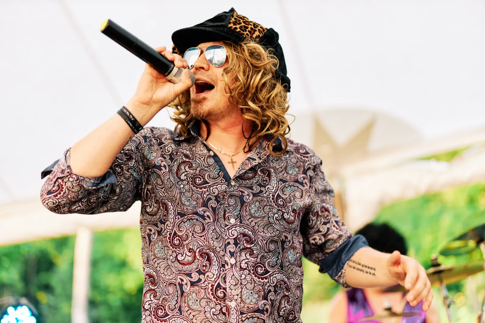 A long-haired band vocalist in a paisley shirt, aviator sunglasses, and a leopard-print cap sings into a handheld microphone under a reception tent. — Tim Larsen Photography, Brainerd Lakes MN