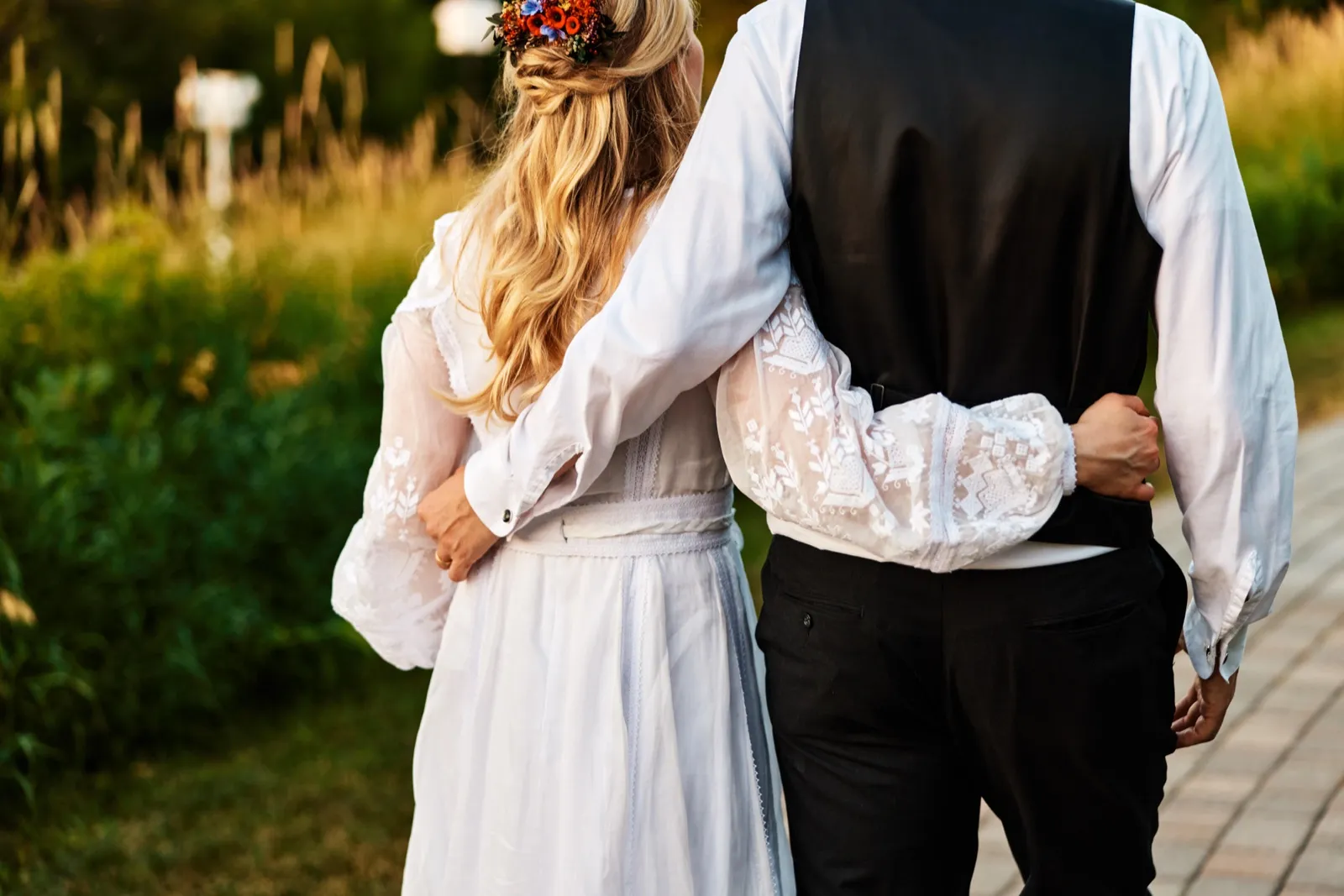 The bride and groom walk away from the camera with arms around each other's backs, her floral crown and embroidered sleeve catching warm evening light along a garden path. — Tim Larsen Photography, Minnesota