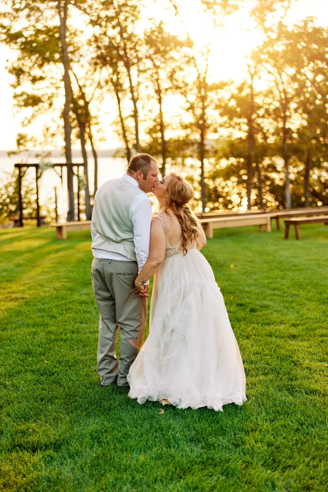 The bride and groom share a kiss on a sunlit lawn beside Gull Lake, with wooden benches and a ceremony arbor catching golden backlight through the trees. — Tim Larsen Photography, Brainerd Lakes MN