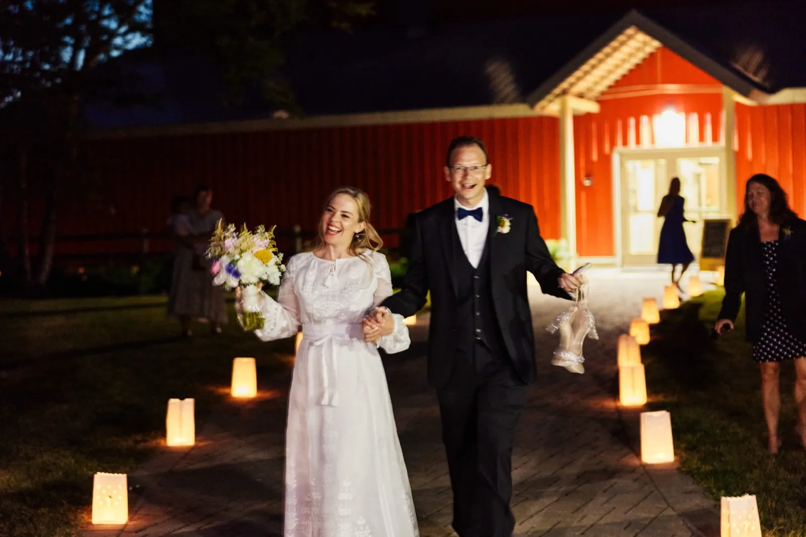 The bride and groom walk hand-in-hand down a candlelit path lined with paper lanterns, with a red barn glowing behind them at night. — Tim Larsen Photography, Minnesota