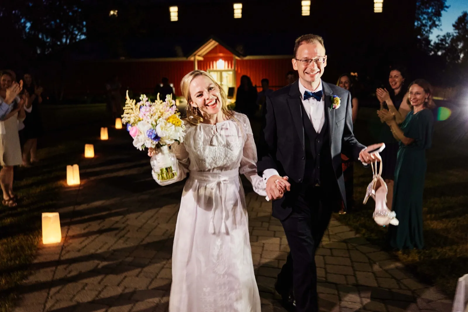 The bride and groom walk hand in hand down a lantern-lit pathway at night while guests applaud on either side, with a red barn glowing behind them. — Tim Larsen Photography, Minnesota