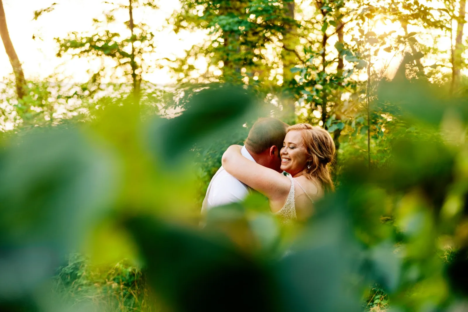 The bride smiles with eyes closed as she embraces the groom in a wooded clearing backlit by golden evening sun, framed by soft green foliage in the foreground. — Tim Larsen Photography, Brainerd Lakes MN