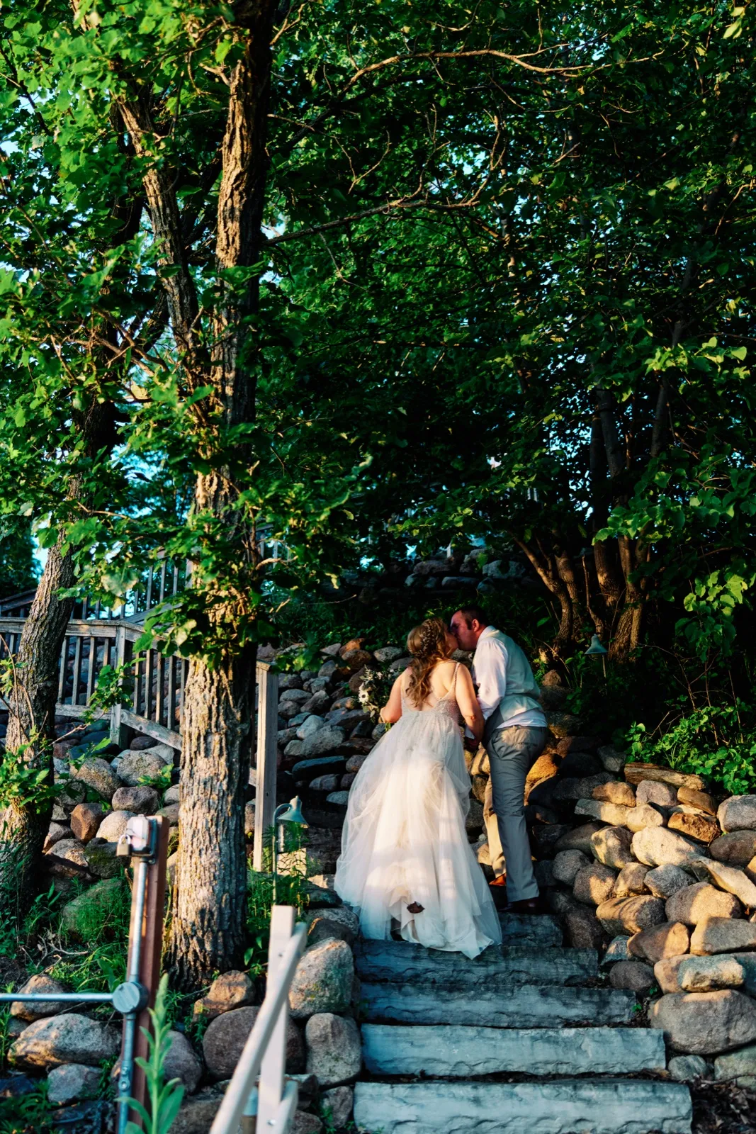 The bride and groom share a kiss while climbing a stone stairway up from the lake, framed by towering oak trees catching warm evening light. — Tim Larsen Photography, Brainerd Lakes MN