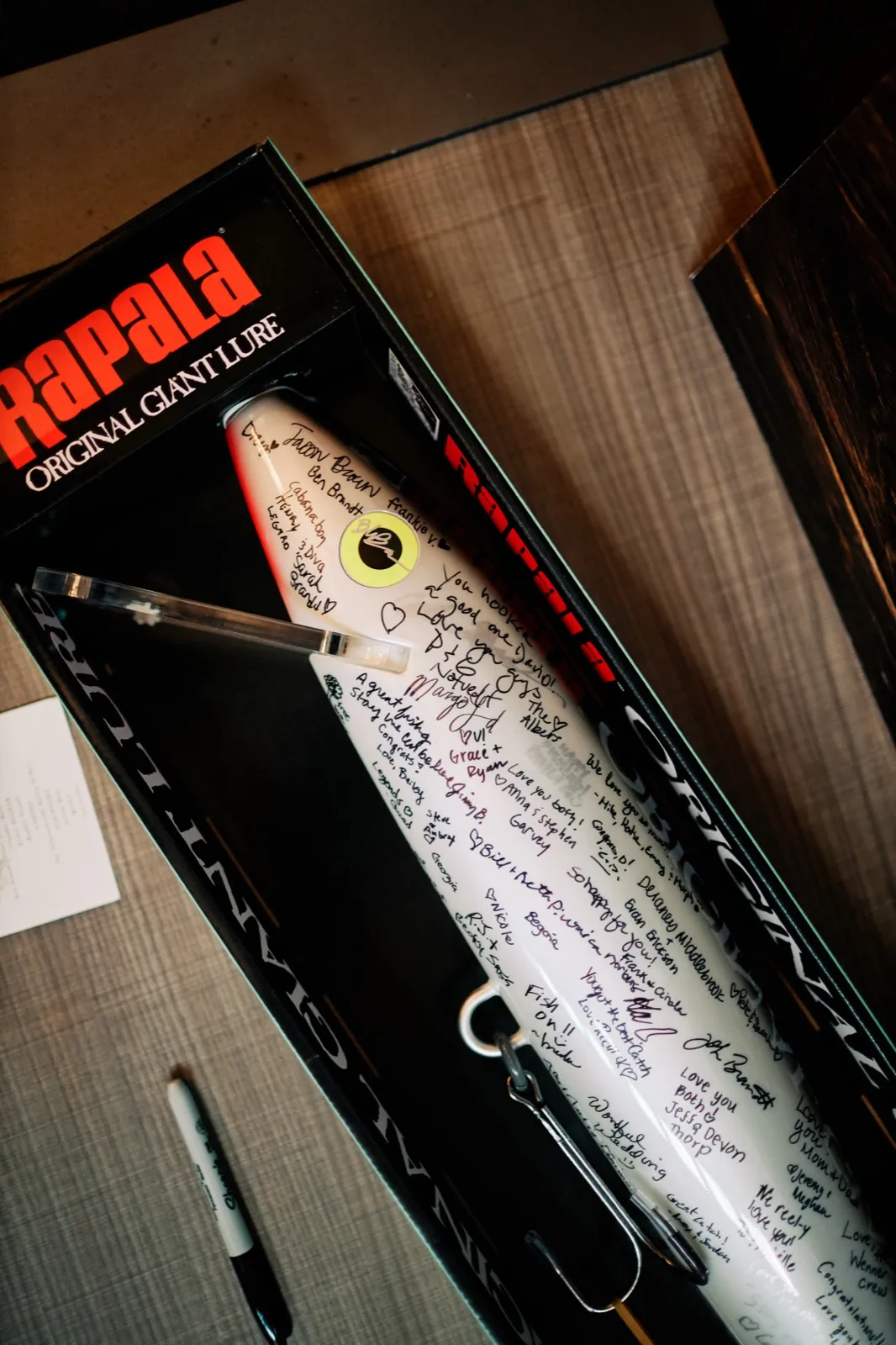 An oversized Rapala Original Giant Lure in its branded box, covered in handwritten signatures and well-wishes from wedding guests, with a marker resting beside it. — Tim Larsen Photography, Brainerd Lakes MN