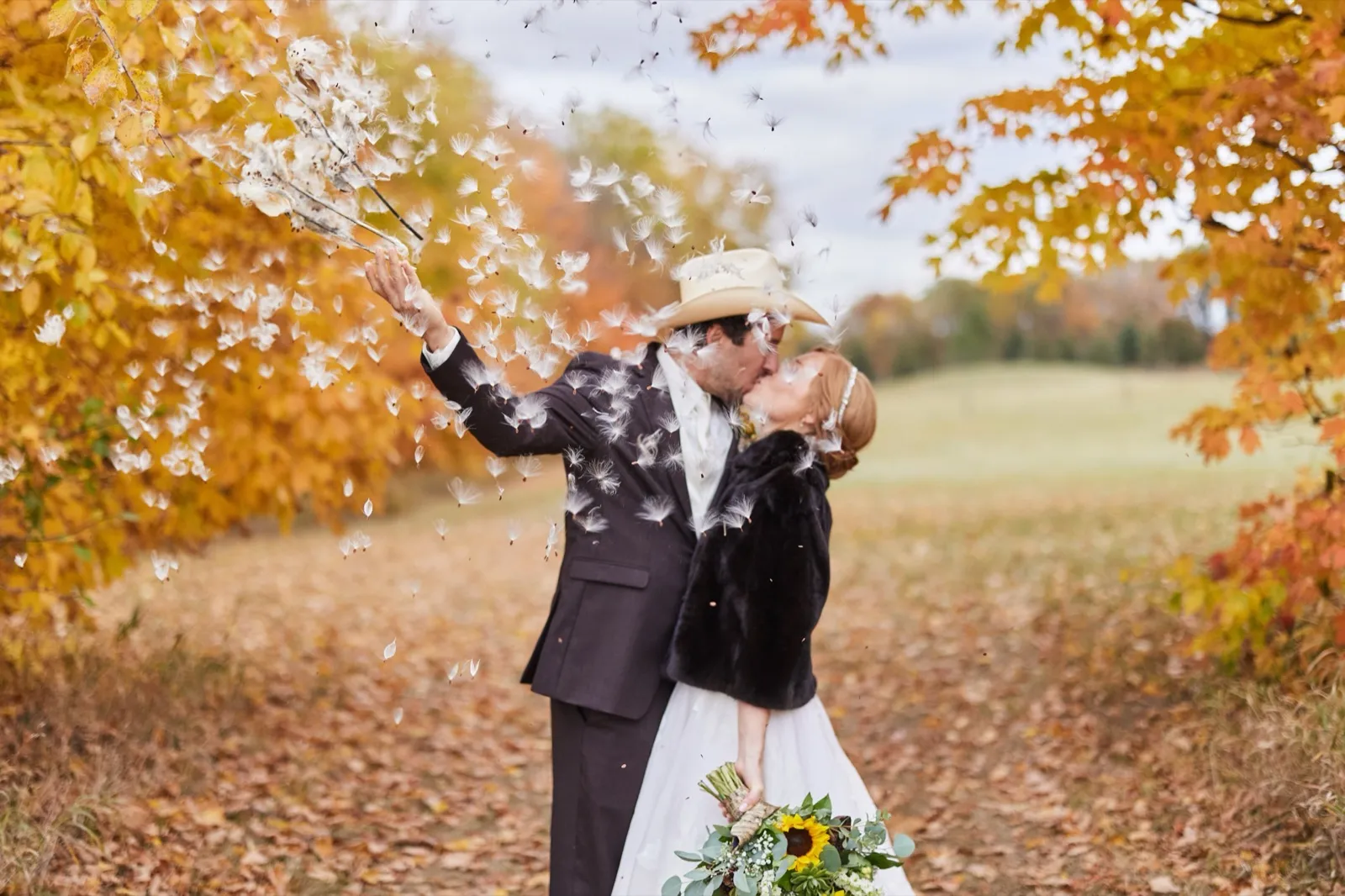 A couple kisses beneath a canopy of bright orange and yellow autumn leaves, tossing seed pods into the air, a sunflower bouquet at the bride's side — Tim Larsen Photography, Brainerd Lakes MN