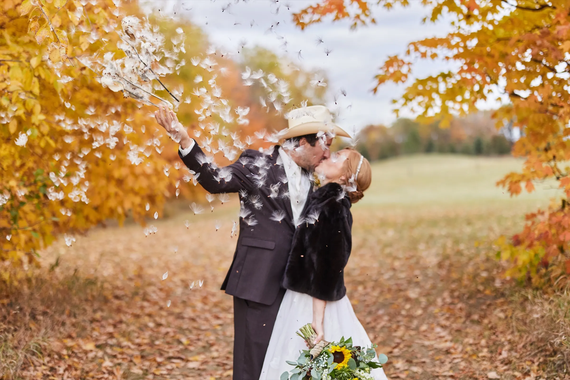 A couple kisses beneath a canopy of bright orange and yellow autumn leaves, tossing seed pods into the air, a sunflower bouquet at the bride's side — Tim Larsen Photography, Brainerd Lakes MN
