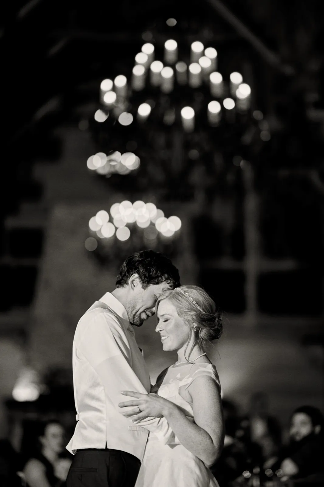 A black-and-white first dance beneath a candle-lit chandelier, the warm glow of the bulbs scattering into soft points of light above the couple — Tim Larsen Photography, Brainerd Lakes MN