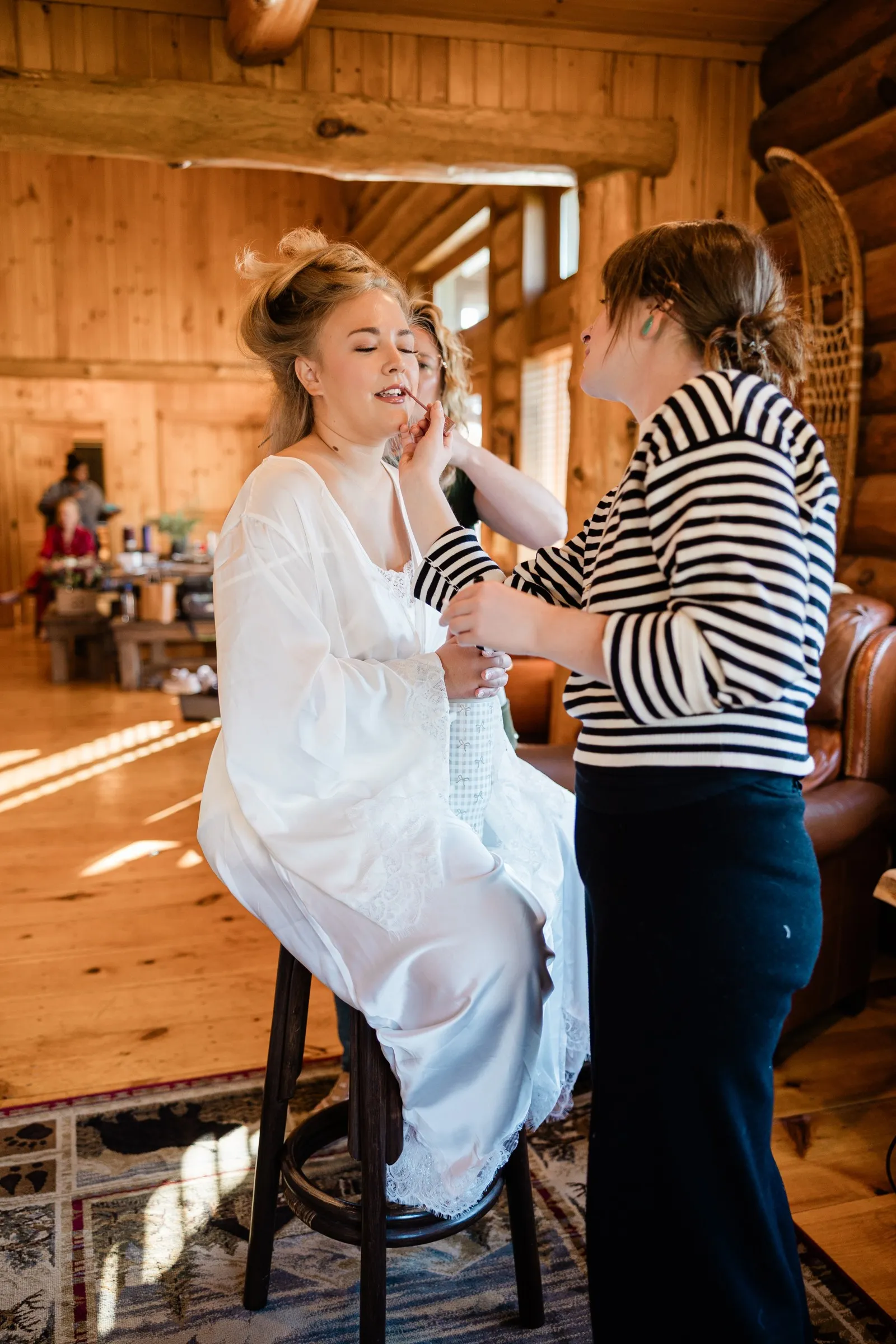 The bride sits in a white robe while a makeup artist applies lip color in a timber room at The Gathering Lodge — Tim Larsen Photography, Brainerd Lakes MN