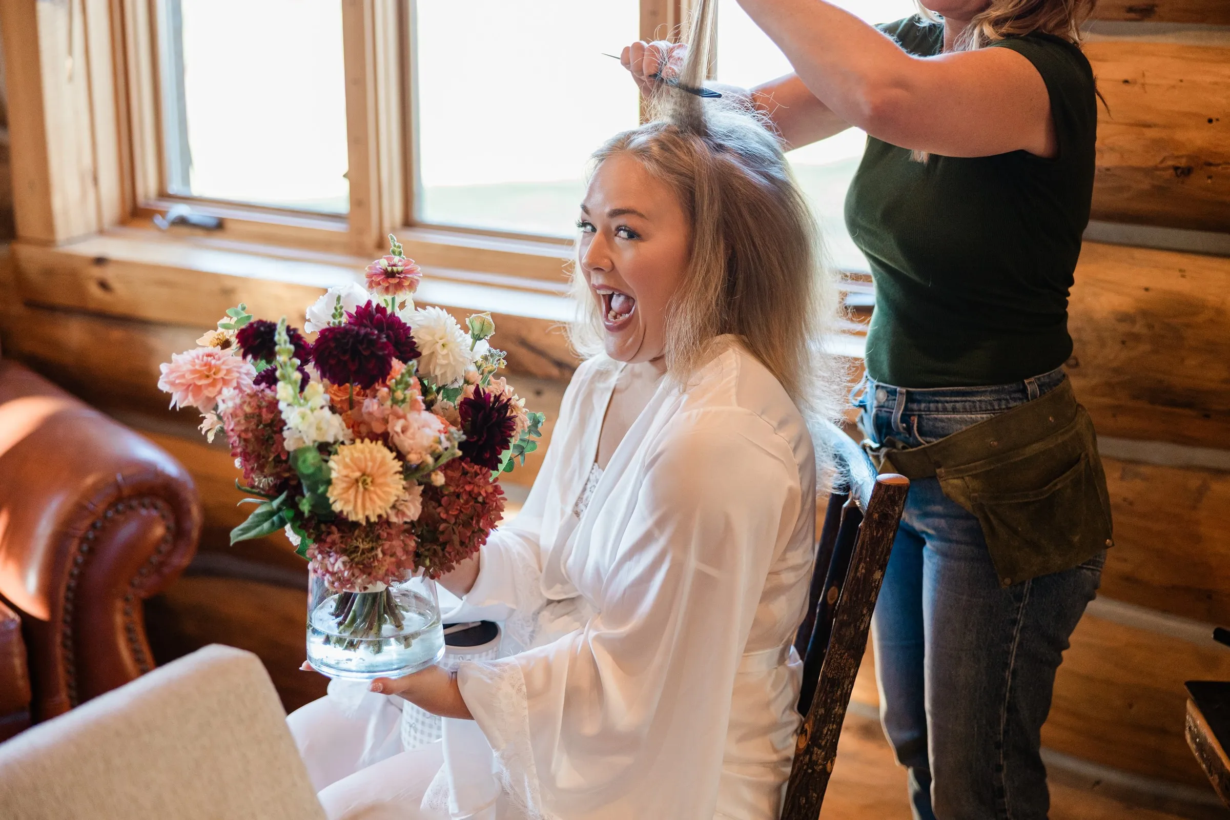 The bride laughs while holding a burgundy, peach and white bouquet as a stylist works on her hair at The Gathering Lodge — Tim Larsen Photography, Brainerd Lakes MN