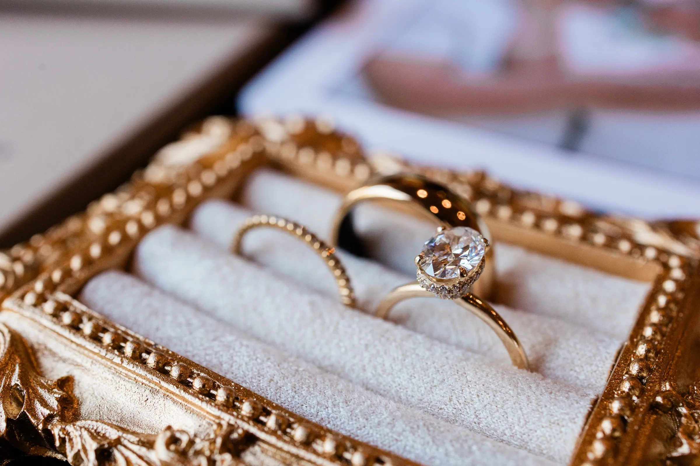 An oval engagement ring sits in front of two wedding bands in a cream-lined gold ring box at The Gathering Lodge — Tim Larsen Photography, Brainerd Lakes MN