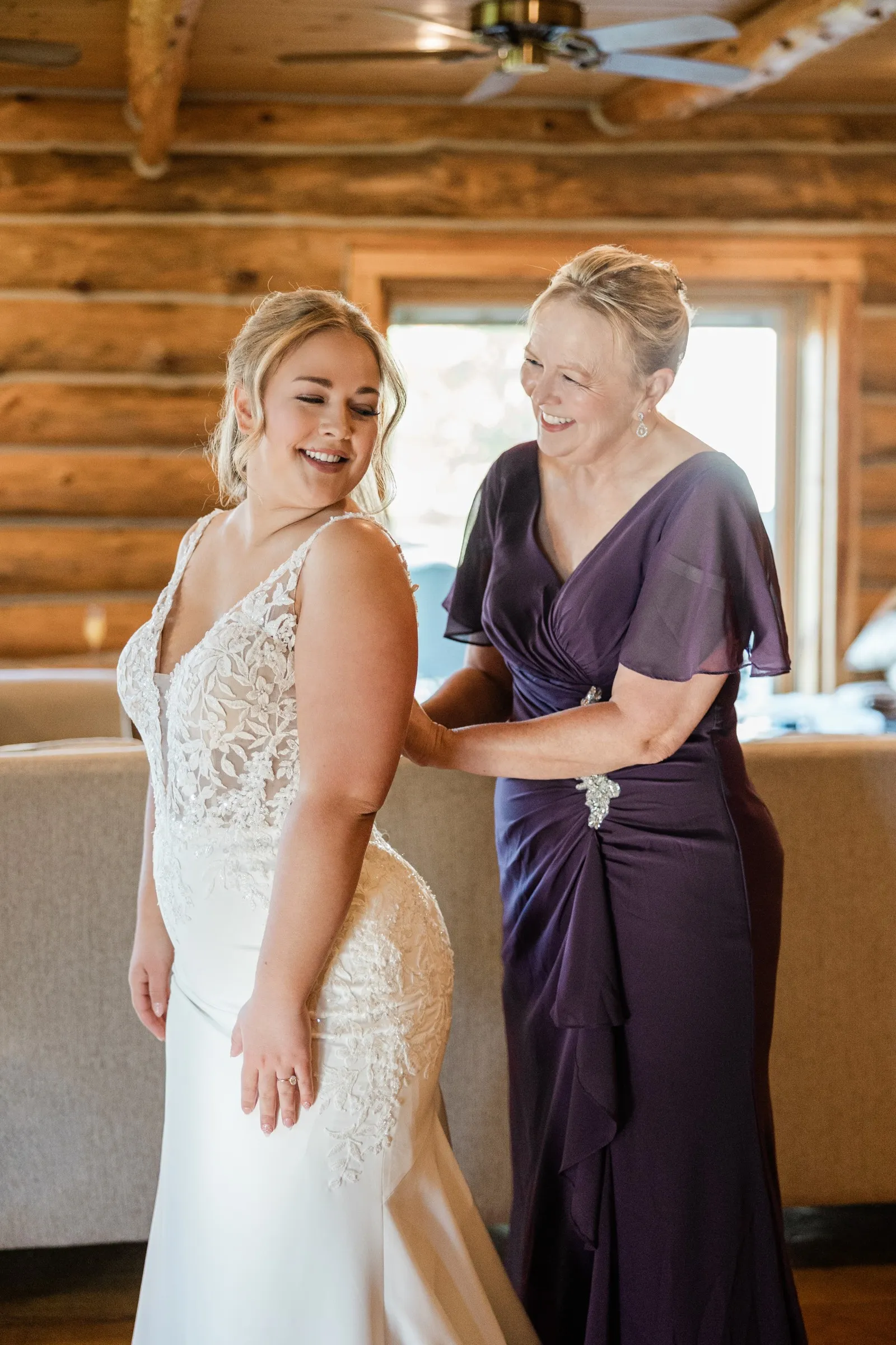 The bride smiles over her shoulder as a woman in a purple dress fastens her gown at The Gathering Lodge — Tim Larsen Photography, Brainerd Lakes MN