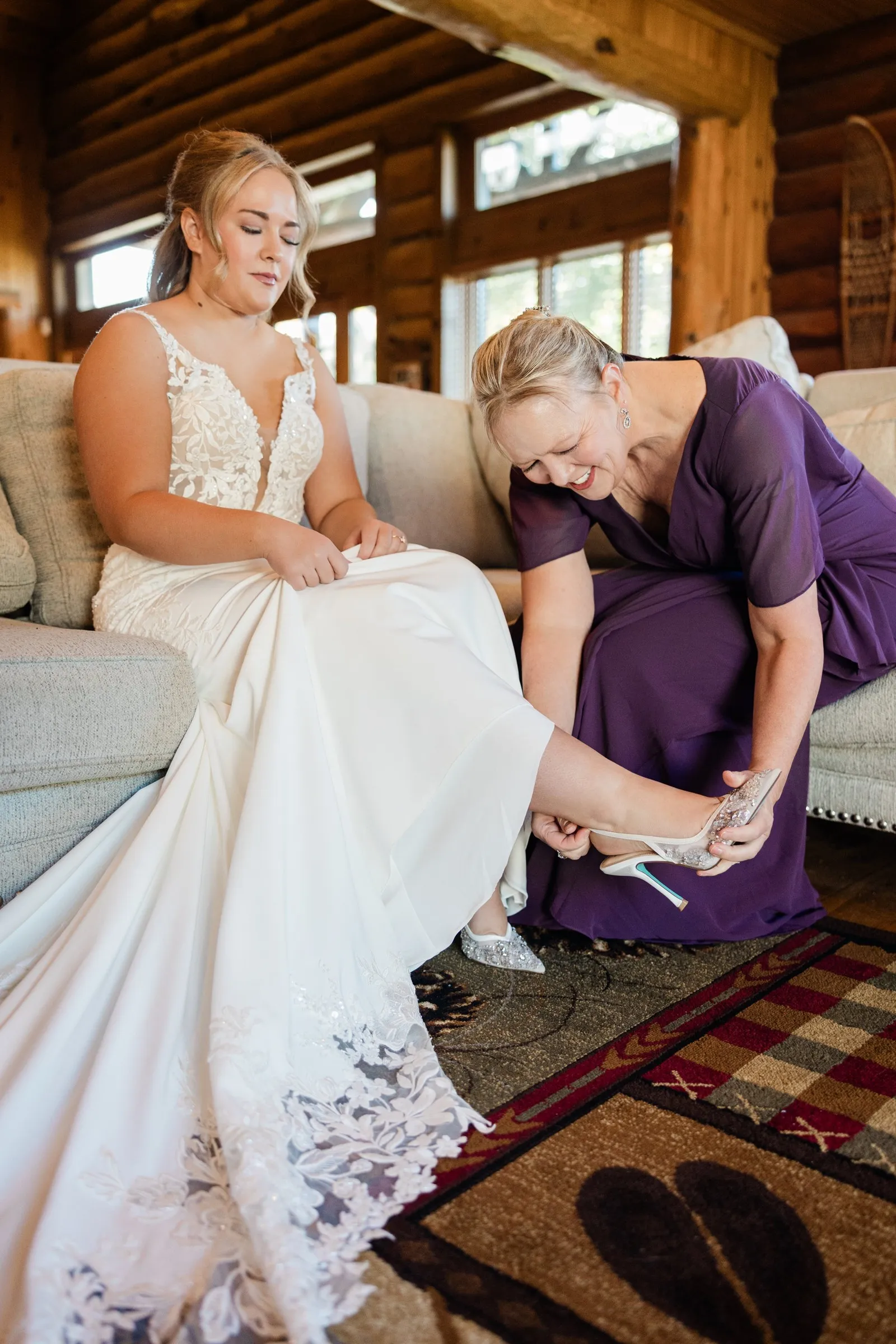 A woman in purple smiles while fastening the bride’s jeweled shoe as the bride sits on a sofa at The Gathering Lodge — Tim Larsen Photography, Brainerd Lakes MN