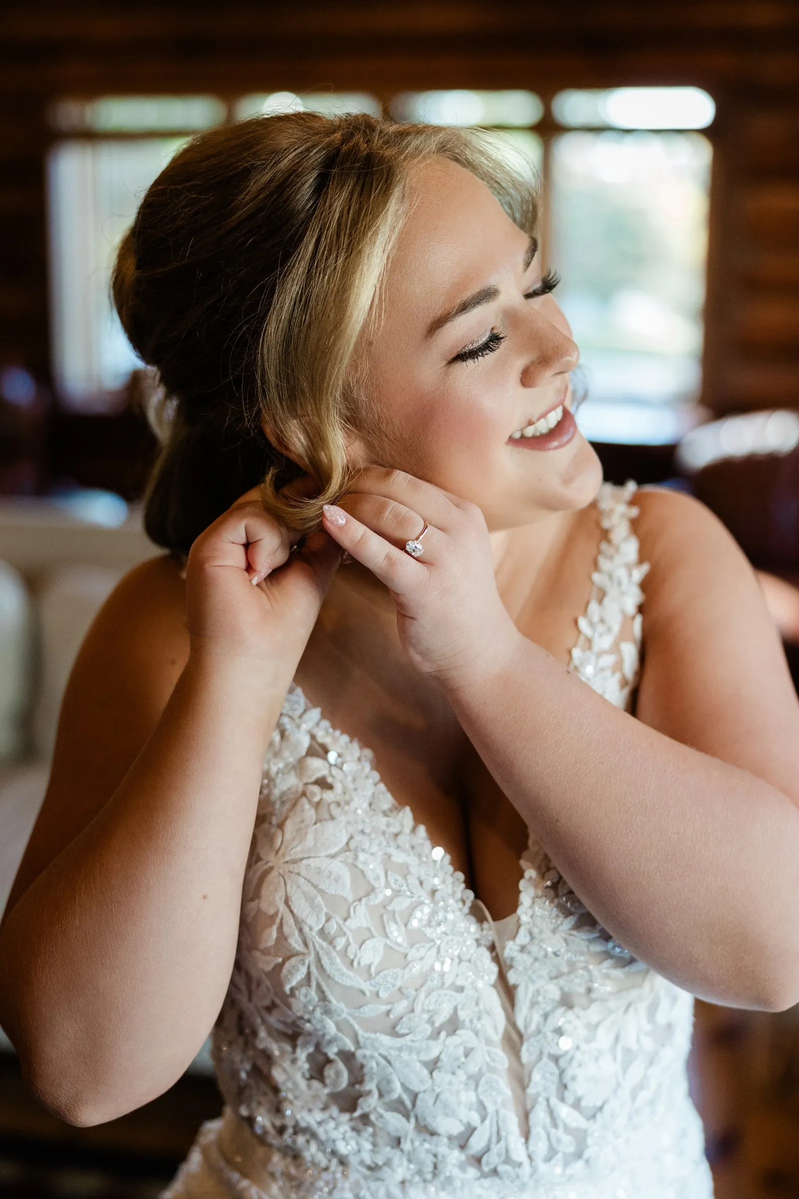 The bride smiles while putting on an earring, with lace on her gown and her engagement ring visible at The Gathering Lodge — Tim Larsen Photography, Brainerd Lakes MN
