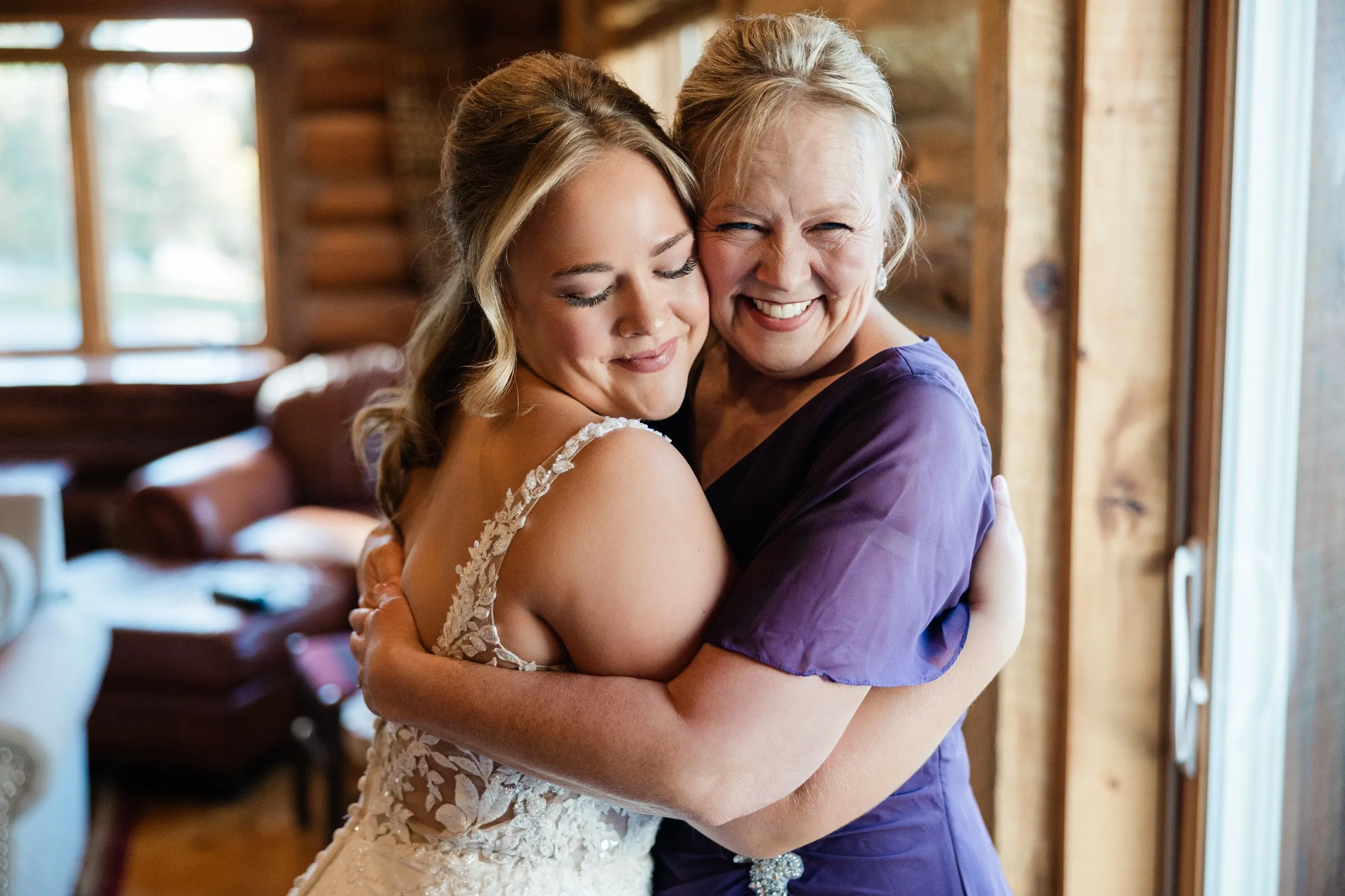 The bride closes her eyes while embracing a smiling woman in purple beside a window at The Gathering Lodge — Tim Larsen Photography, Brainerd Lakes MN