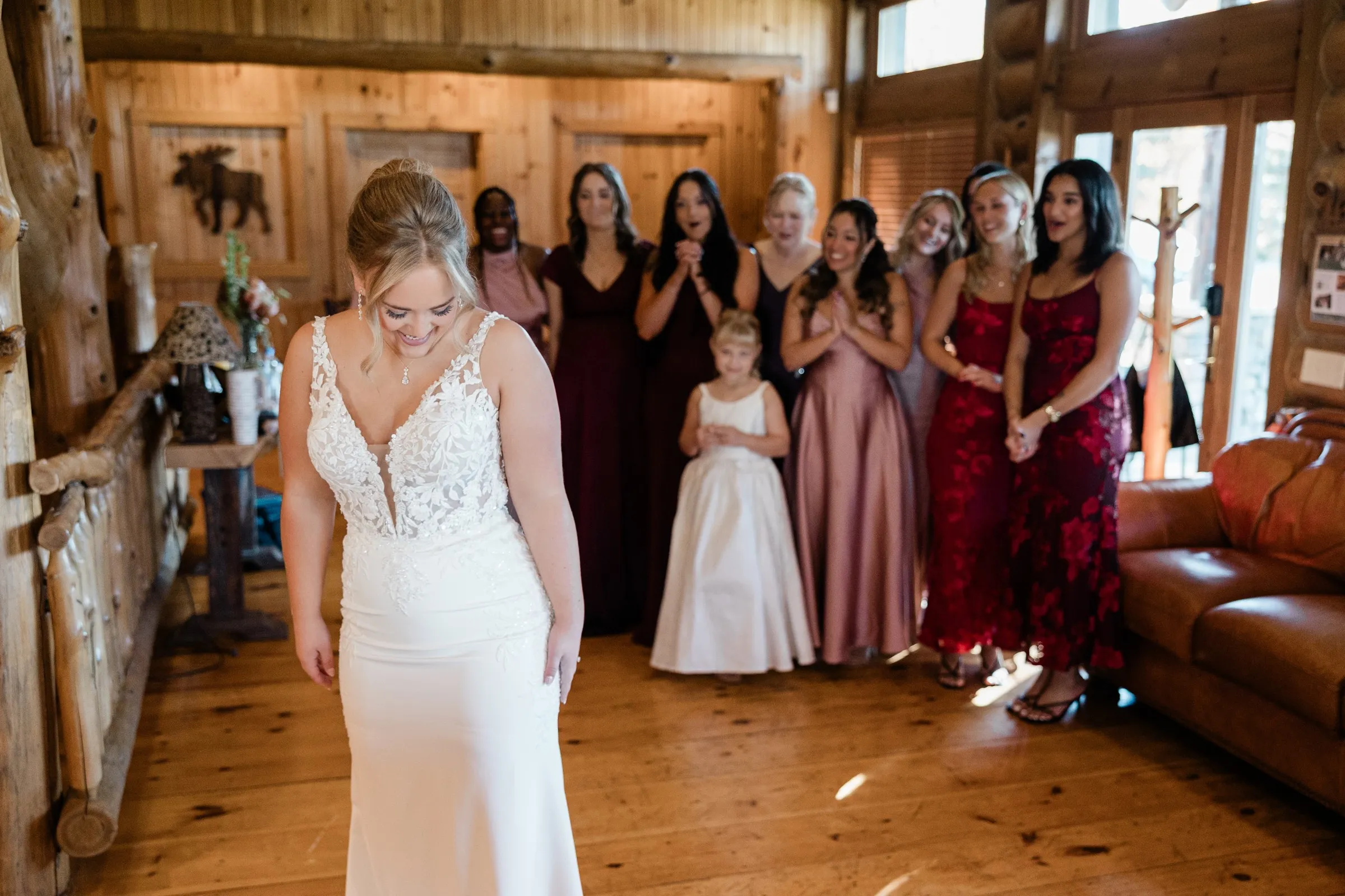 The bride smiles down at her gown while attendants watch and applaud behind her at The Gathering Lodge — Tim Larsen Photography, Brainerd Lakes MN