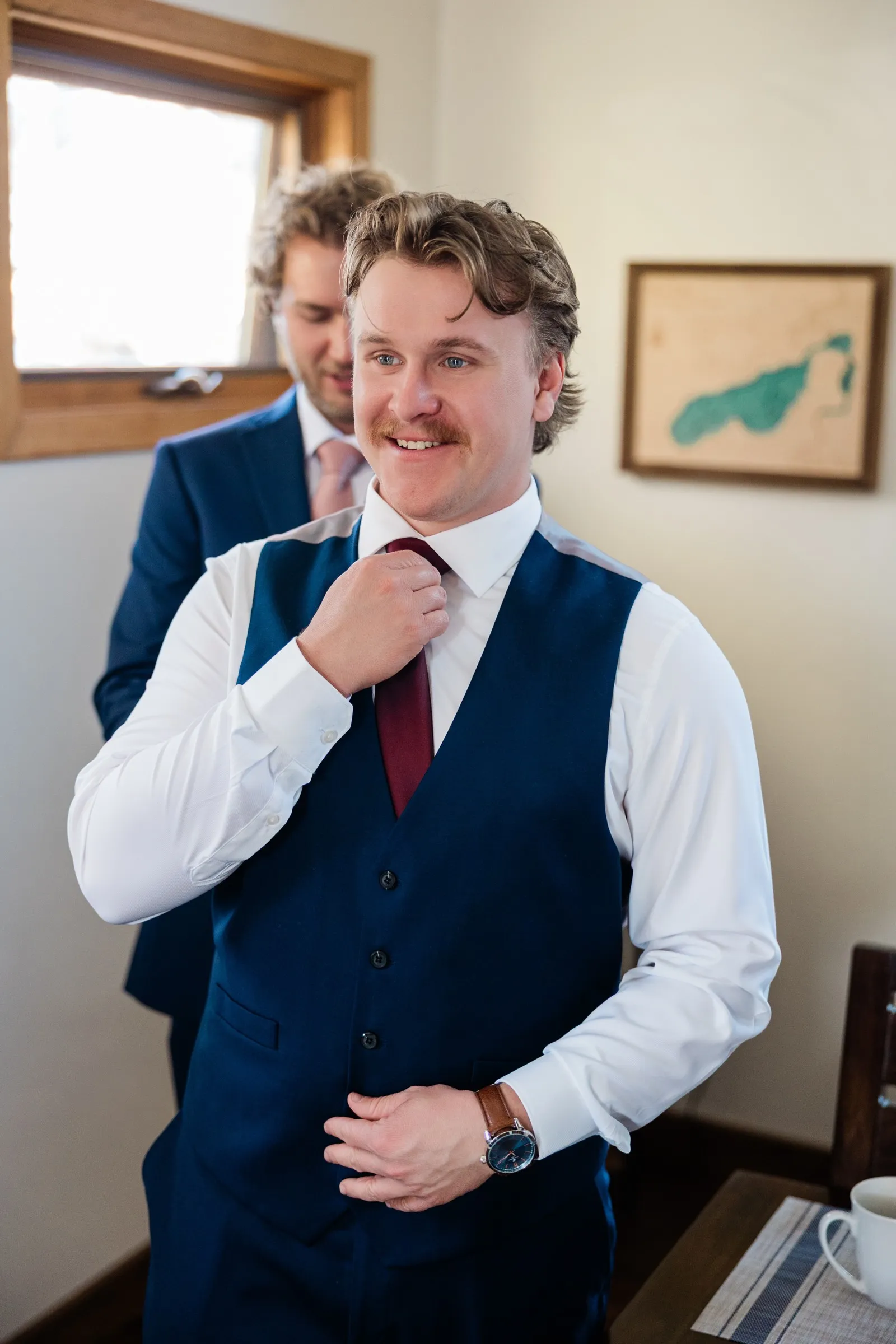 The groom adjusts his burgundy tie while another man in a blue suit stands behind him — Tim Larsen Photography, Brainerd Lakes MN