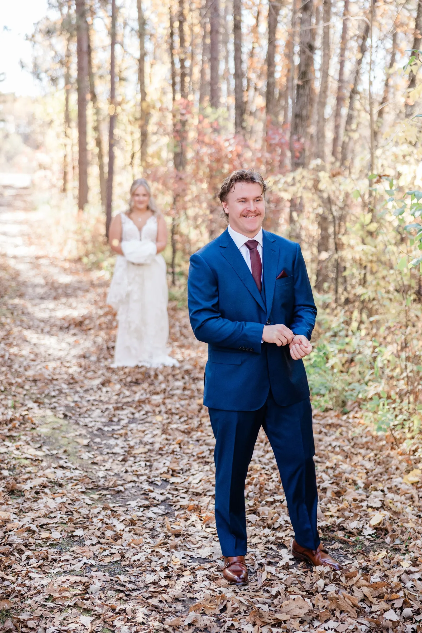 The groom adjusts his sleeve in the foreground while the bride waits behind him on a wooded path — Tim Larsen Photography, Brainerd Lakes MN