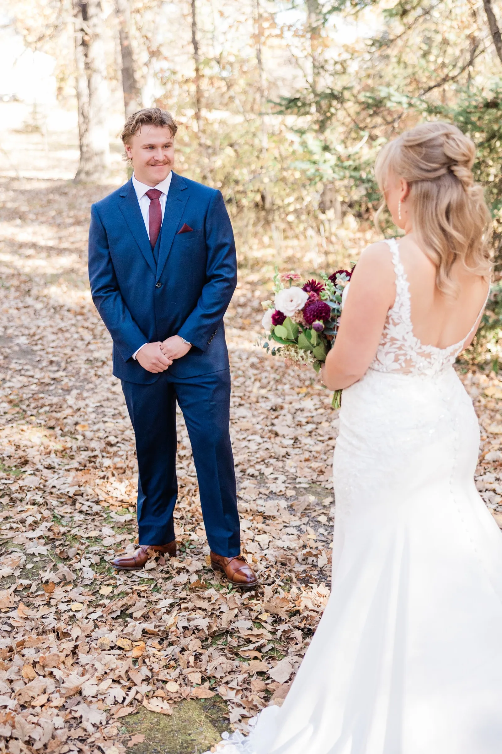 The groom smiles at the bride across the leaf-covered path as she faces him with her bouquet — Tim Larsen Photography, Brainerd Lakes MN