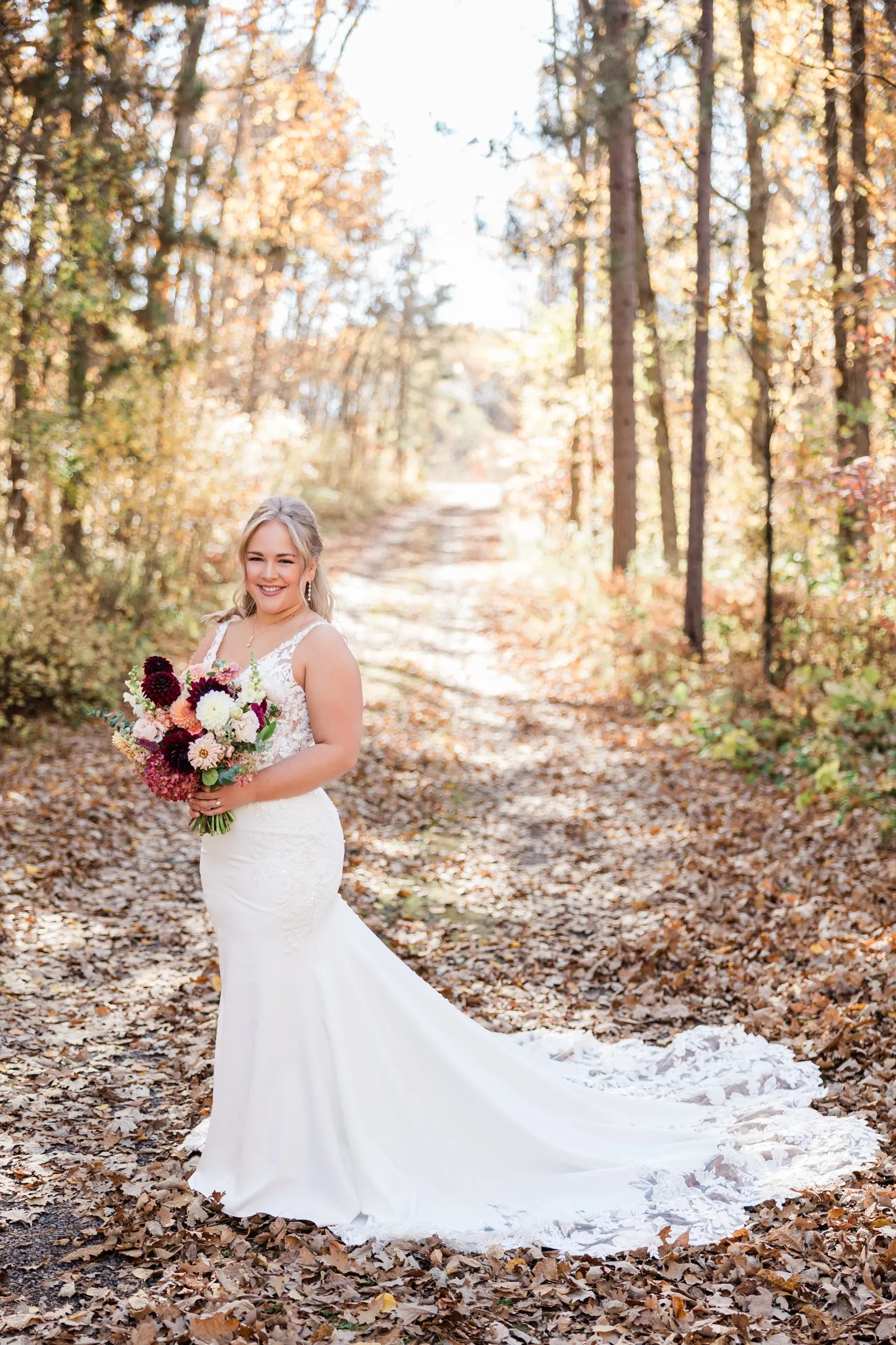 The bride holds her bouquet with her lace train spread across fallen leaves on a wooded path — Tim Larsen Photography, Brainerd Lakes MN