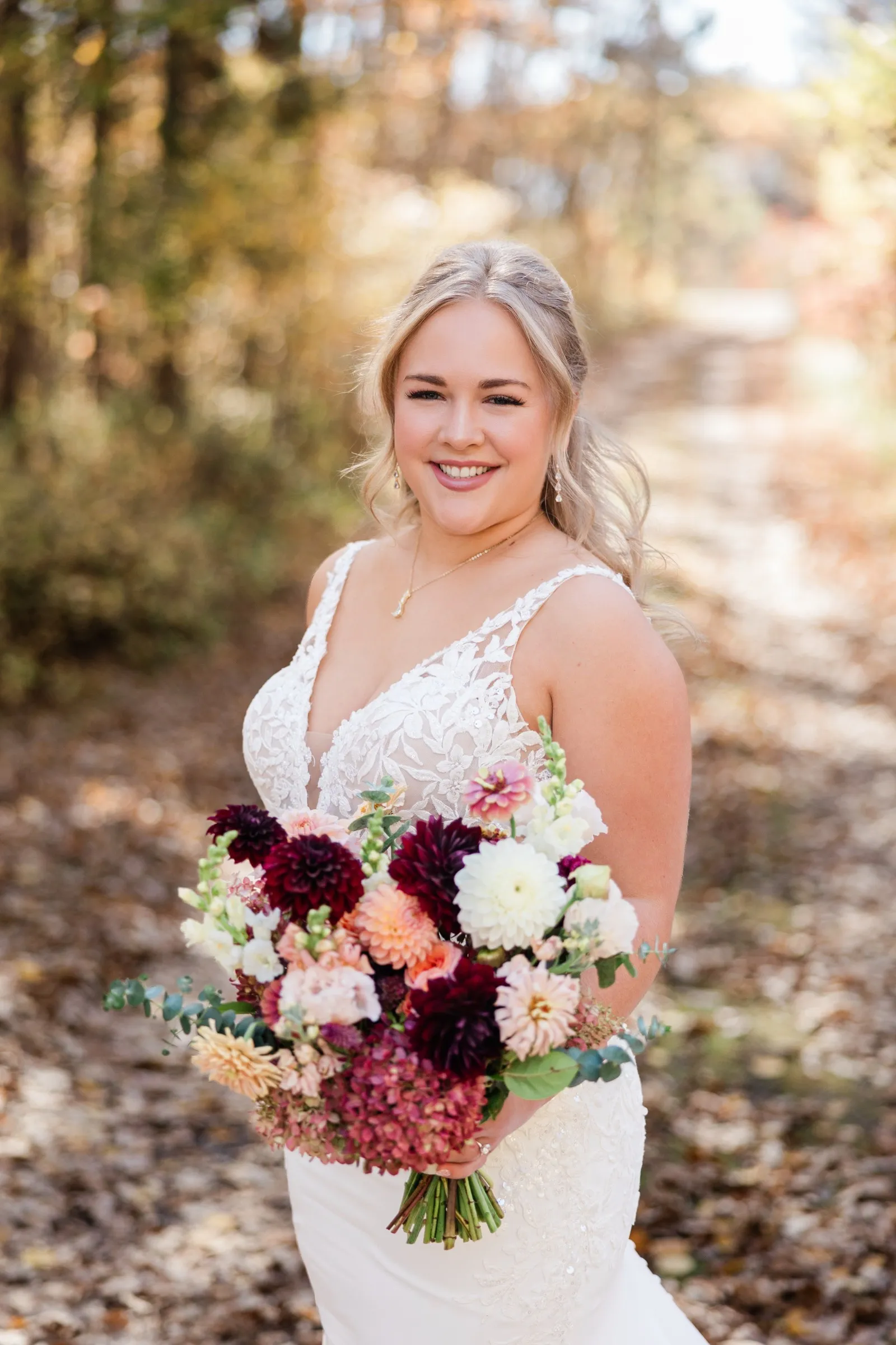 The bride smiles while holding a bouquet of burgundy, blush, peach, and white flowers in the woods — Tim Larsen Photography, Brainerd Lakes MN