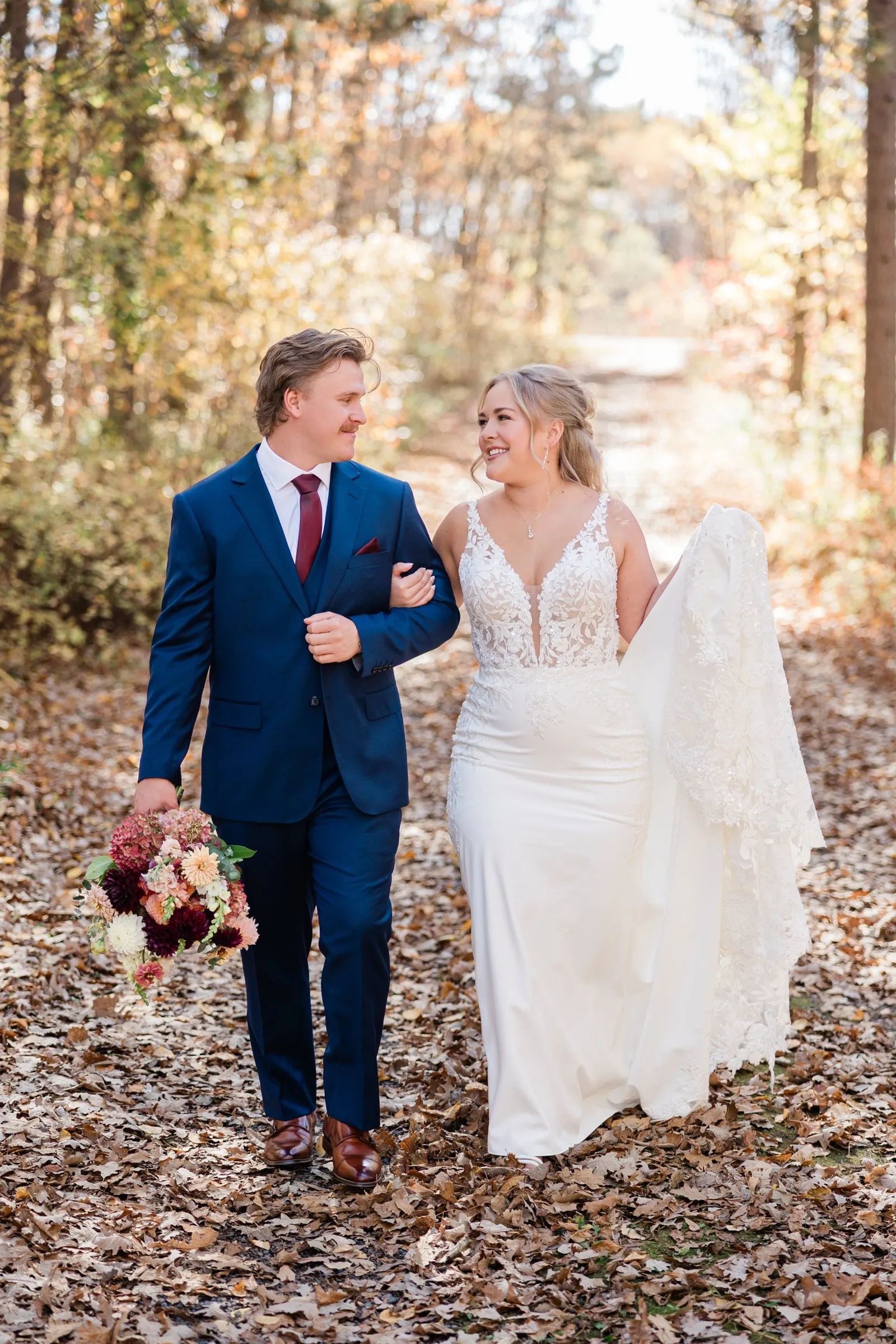 The couple walk arm in arm through fallen leaves as the groom carries the bouquet and the bride lifts her train — Tim Larsen Photography, Brainerd Lakes MN