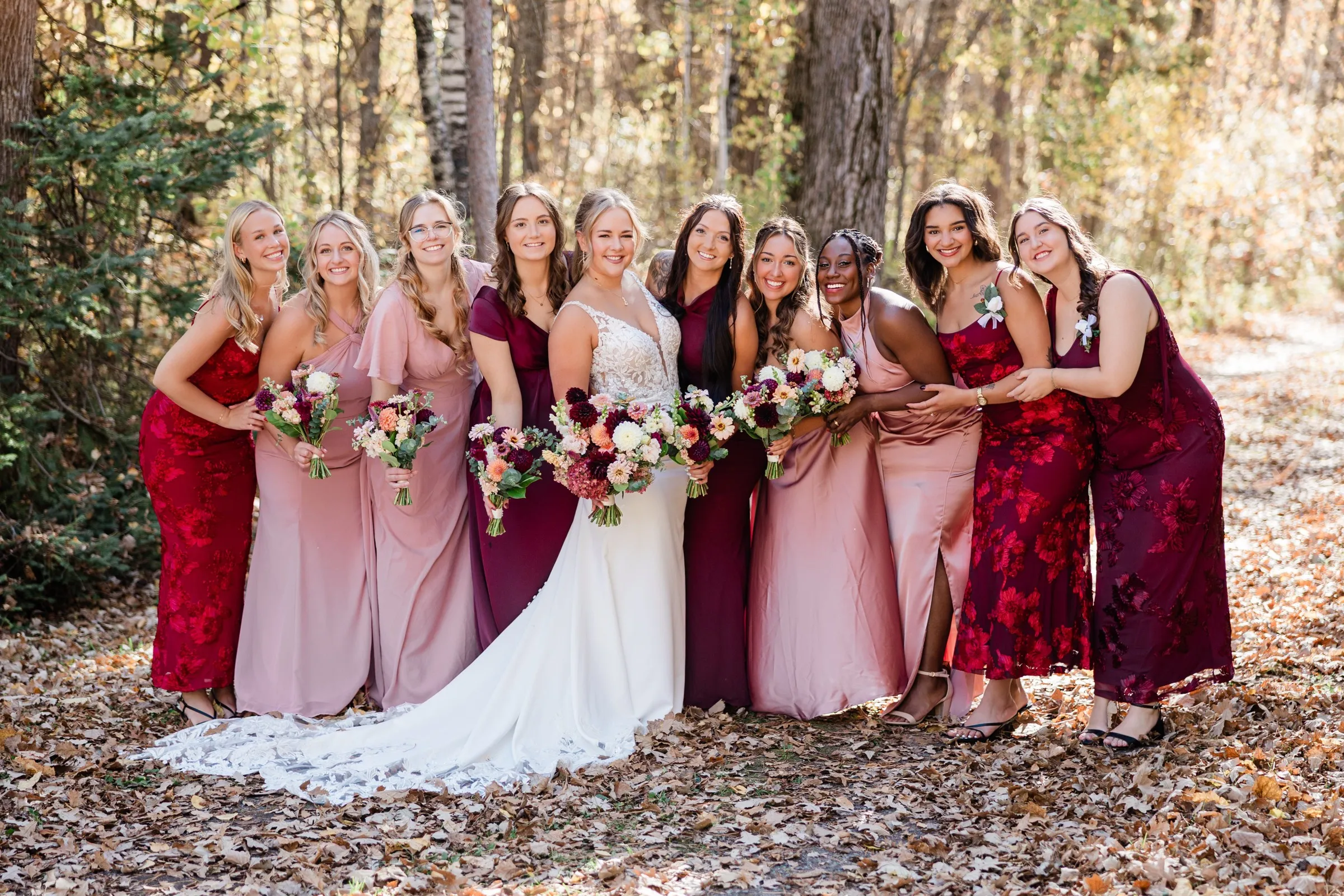 The bride stands with nine women in burgundy and blush dresses on a leaf-covered path — Tim Larsen Photography, Brainerd Lakes MN