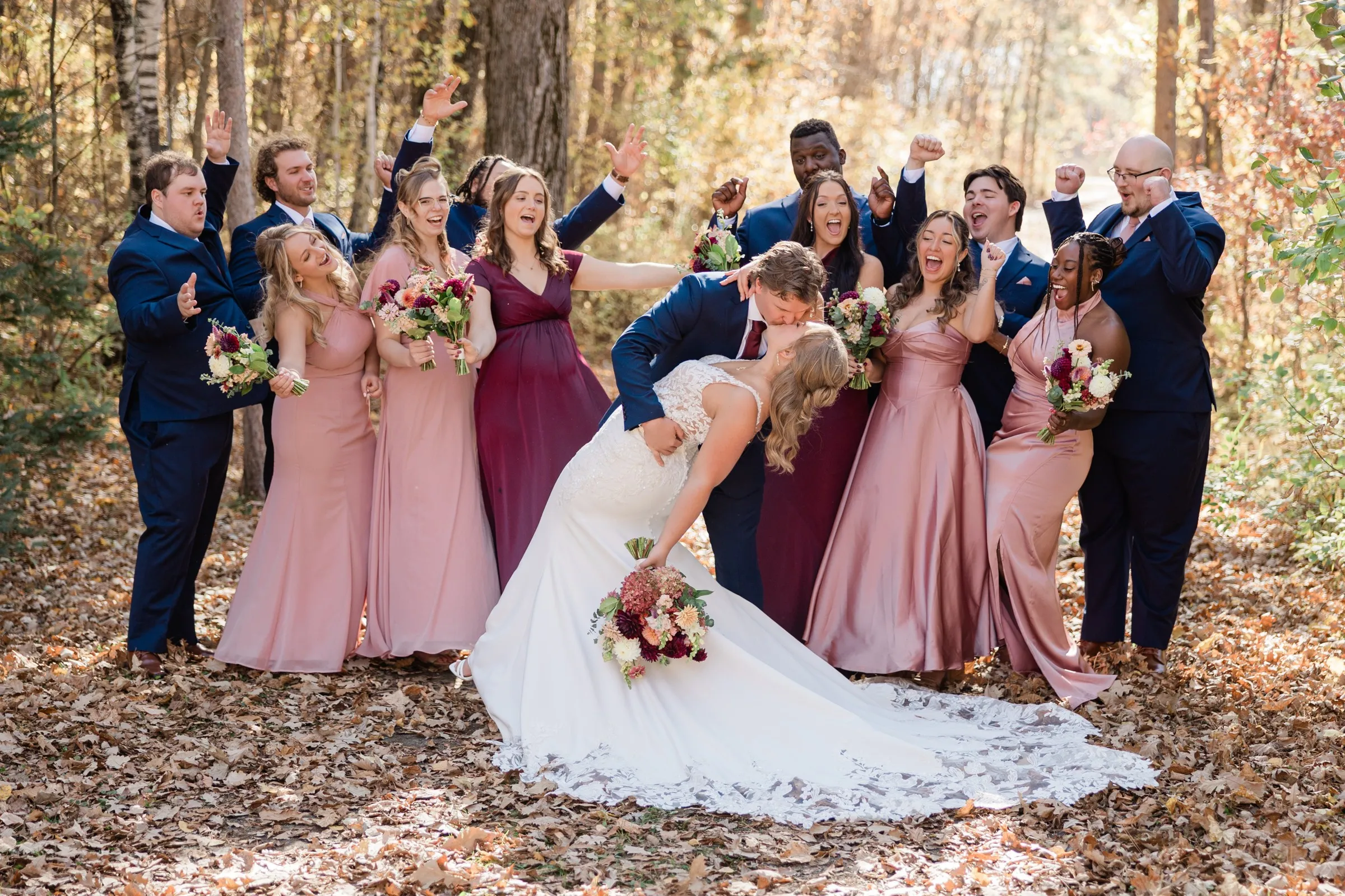 The groom dips and kisses the bride while their wedding party cheers with raised arms among autumn trees — Tim Larsen Photography, Brainerd Lakes MN
