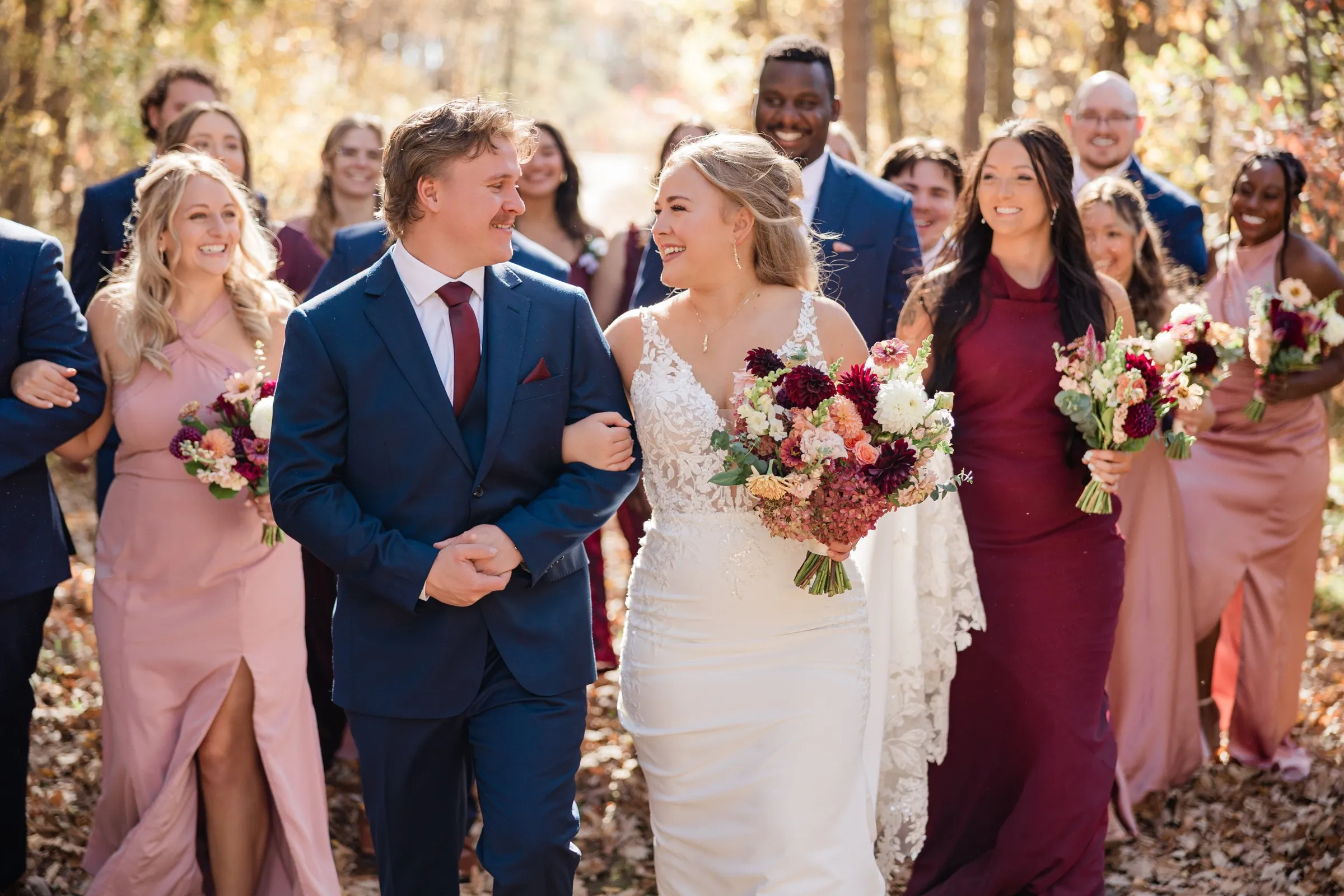 The couple smile at each other while walking arm in arm ahead of their wedding party on a wooded path — Tim Larsen Photography, Brainerd Lakes MN