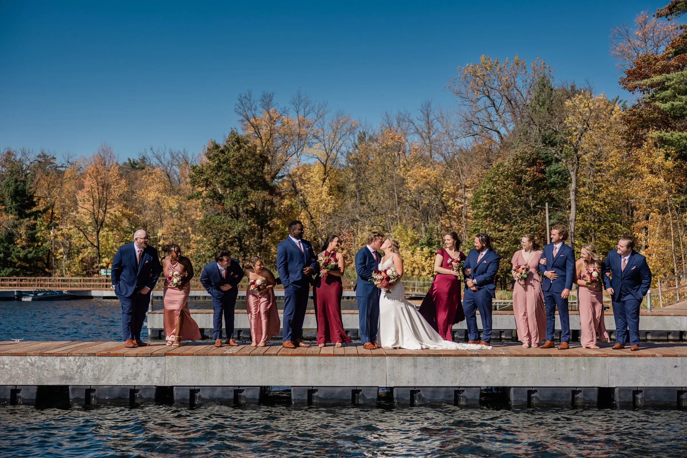 The couple kiss at the center of their wedding party spread across a dock with autumn trees and blue water behind them — Tim Larsen Photography, Brainerd Lakes MN