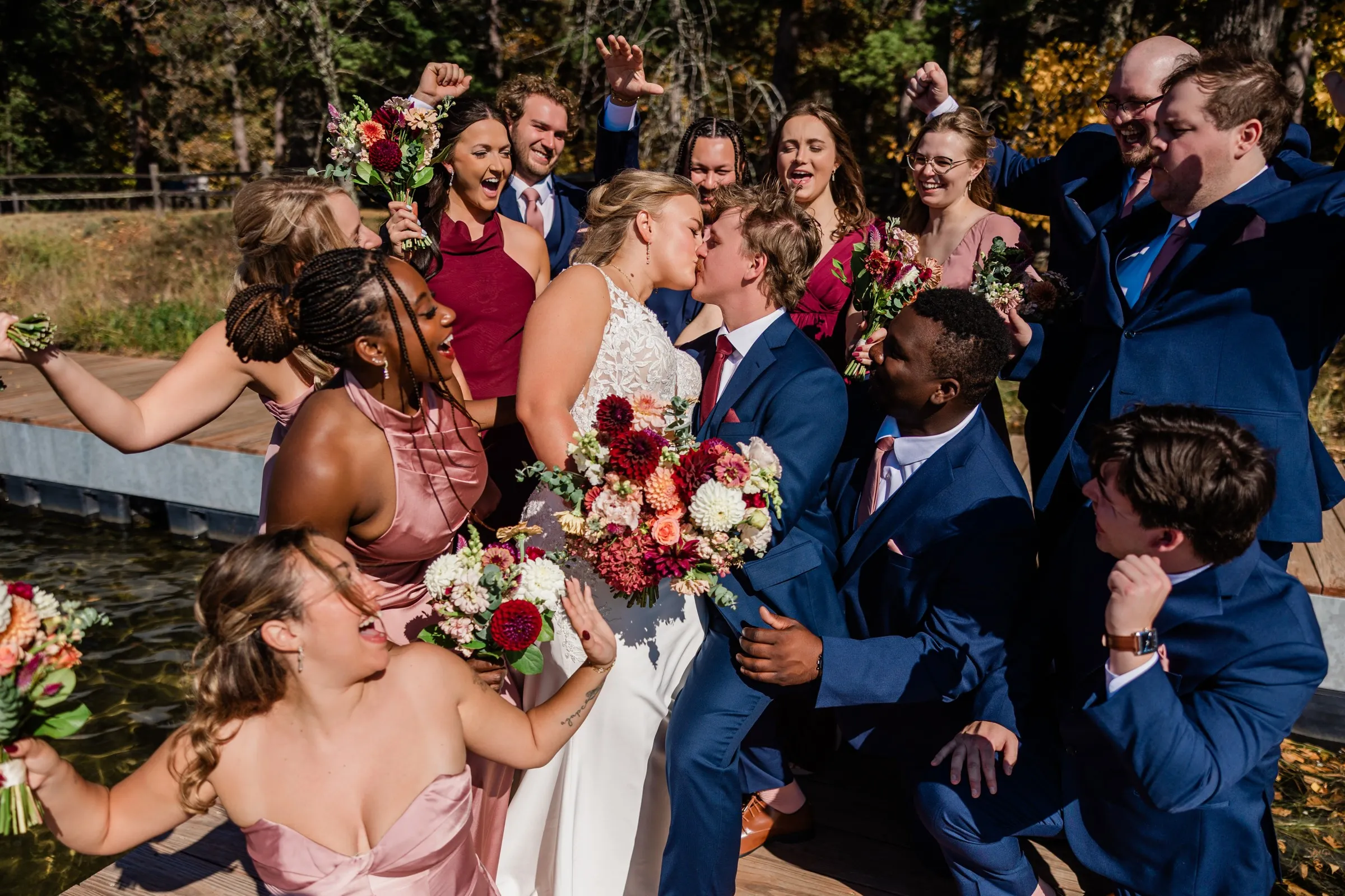 The couple kiss on the dock while attendants crowd around them cheering with bouquets and raised arms — Tim Larsen Photography, Brainerd Lakes MN