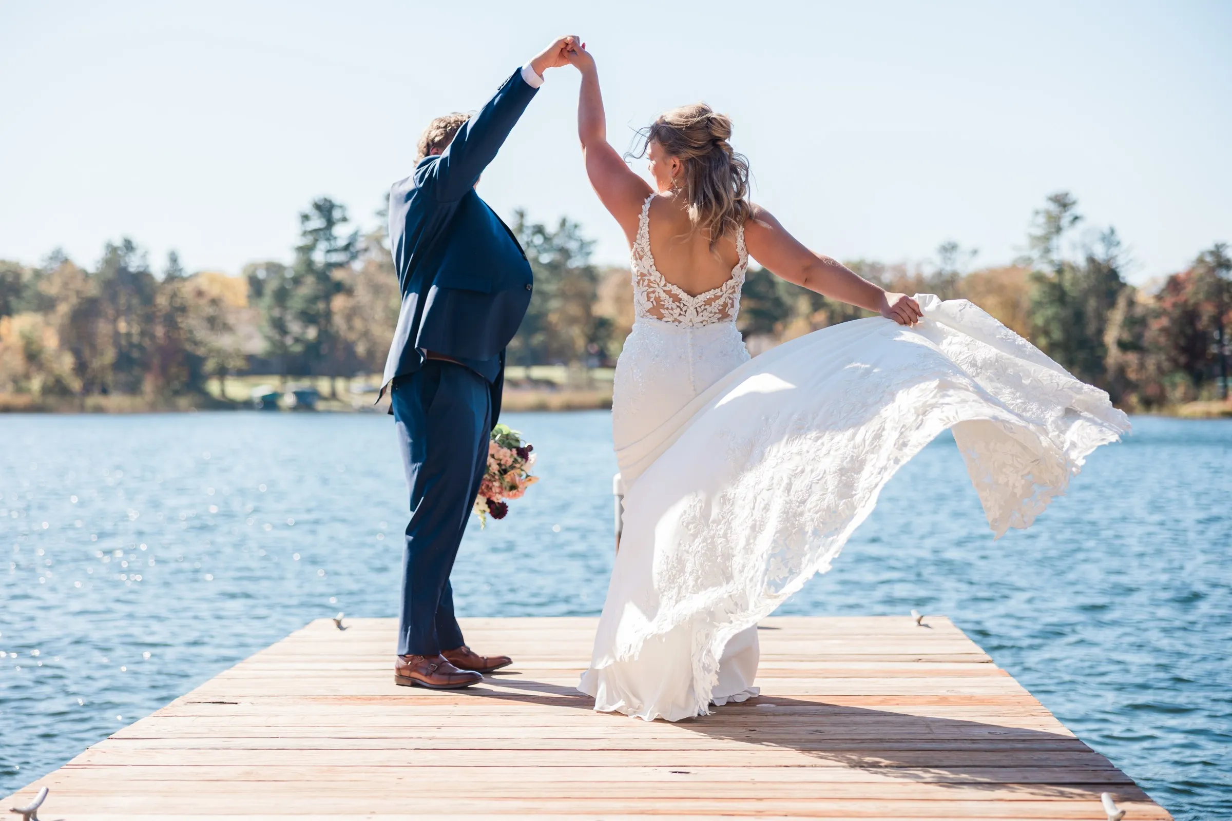 The groom raises their joined hands as the bride twirls on the dock, holding her lace train out above the water — Tim Larsen Photography, Brainerd Lakes MN