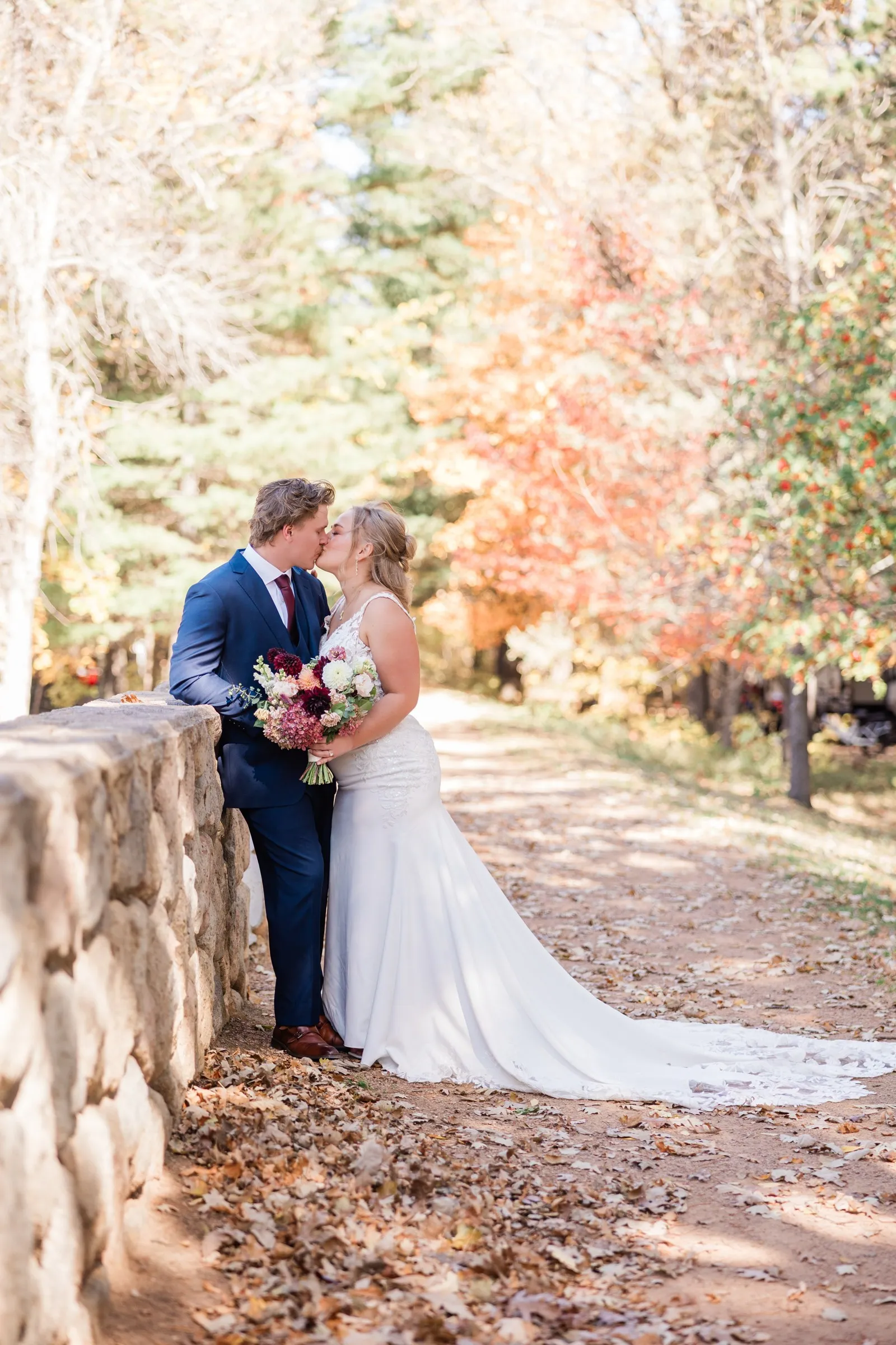 The couple kiss beside a stone wall with the bride's lace train spread over a path beneath autumn trees — Tim Larsen Photography, Brainerd Lakes MN