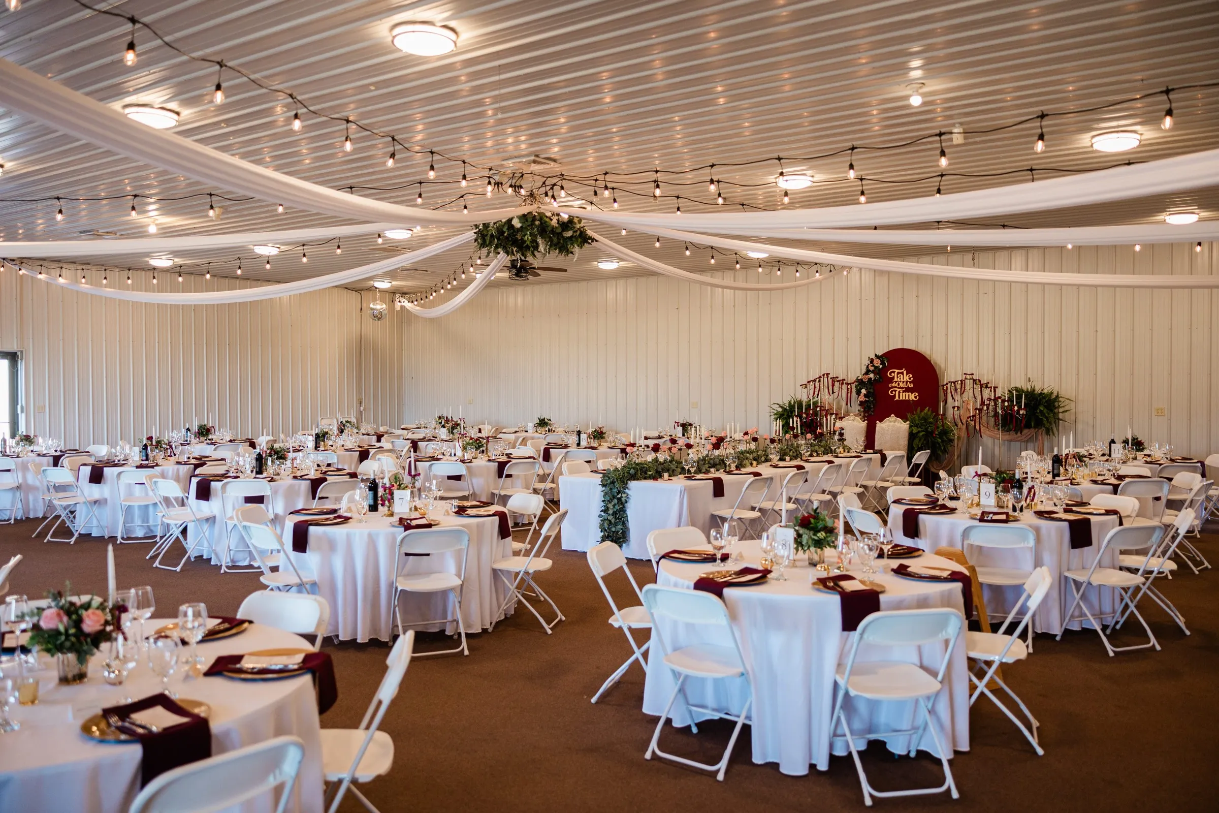 The reception room has white tables, burgundy napkins, ceiling drapery, and string lights beneath a floral centerpiece — Tim Larsen Photography, Brainerd Lakes MN