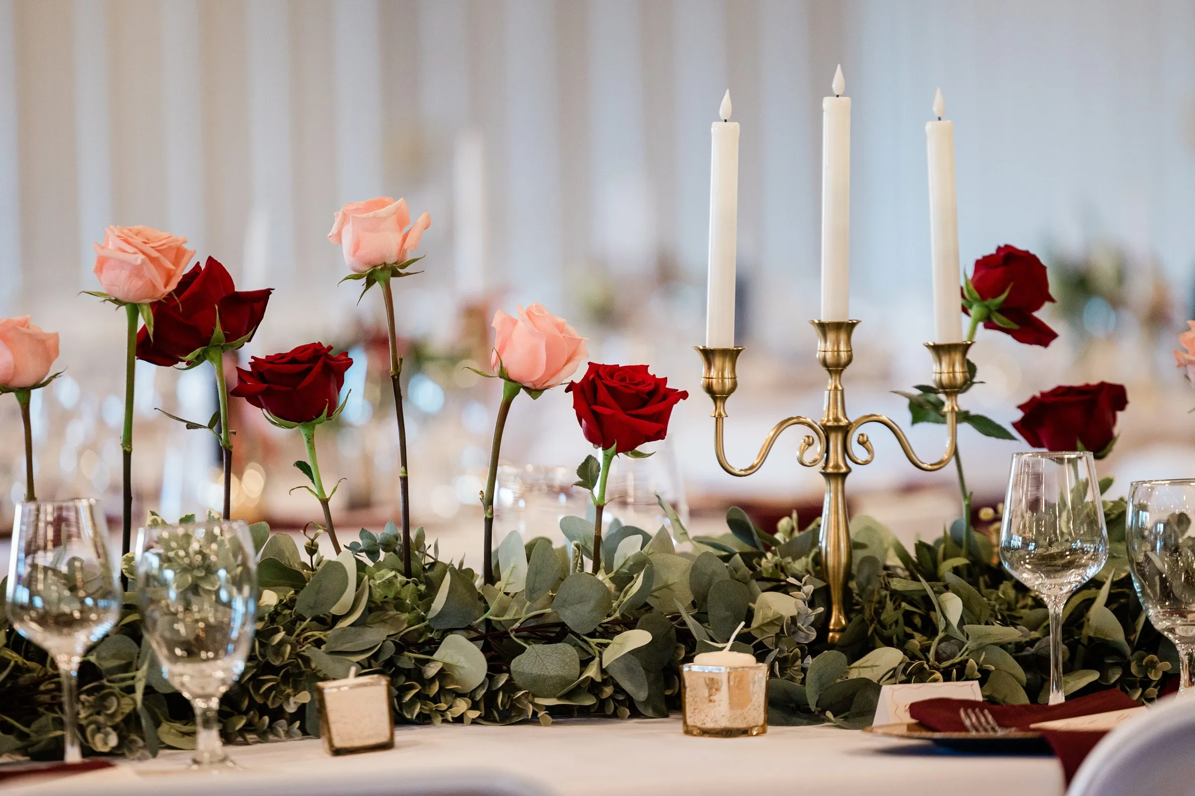 Red and blush roses rise from a greenery runner beside a gold candleholder, wine glasses, and burgundy napkins — Tim Larsen Photography, Brainerd Lakes MN