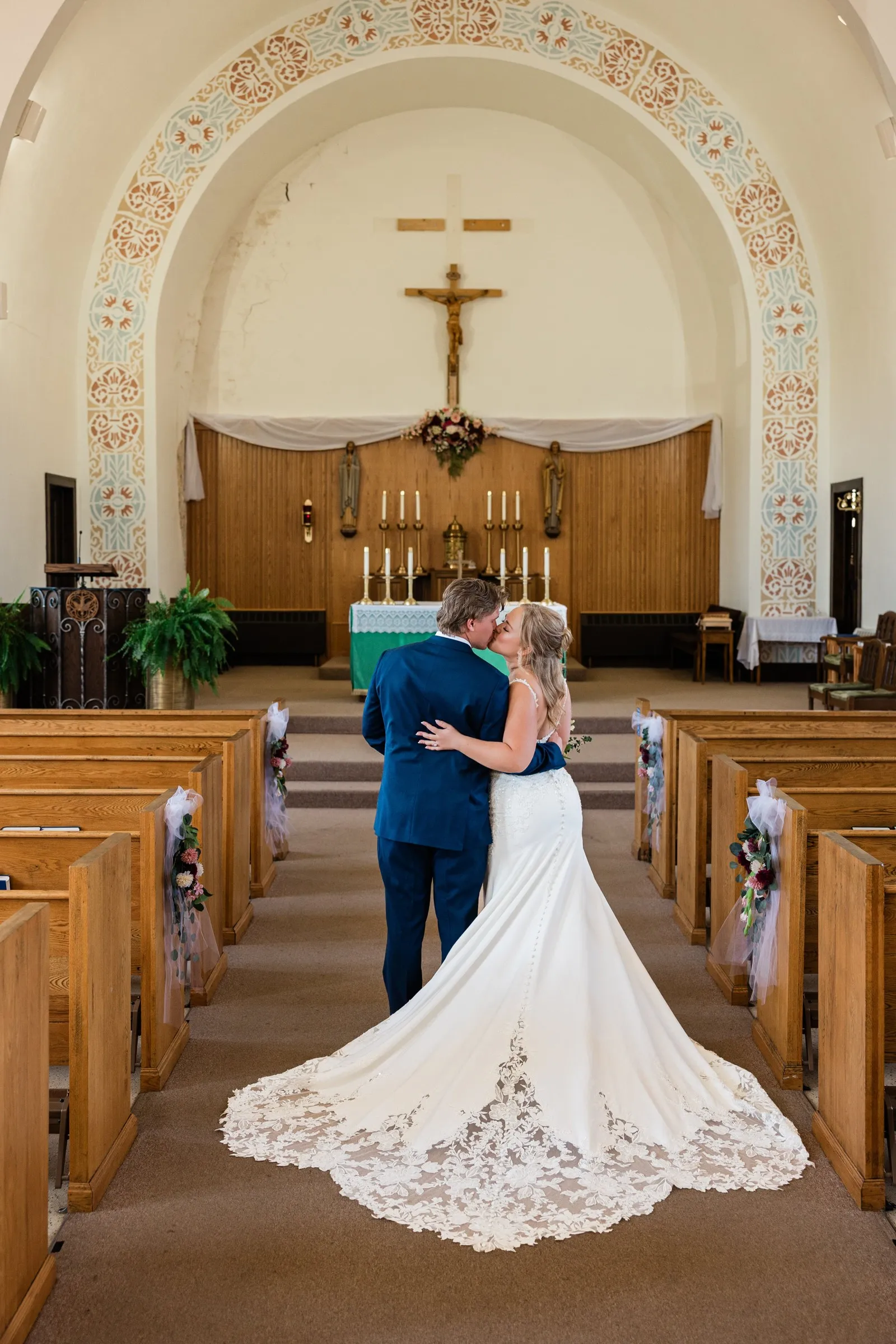 The couple kiss in an empty church aisle beneath a decorated arch, with the bride's lace train spread between the pews at St. Joseph Catholic Church in Crosby — Tim Larsen Photography, Brainerd Lakes MN
