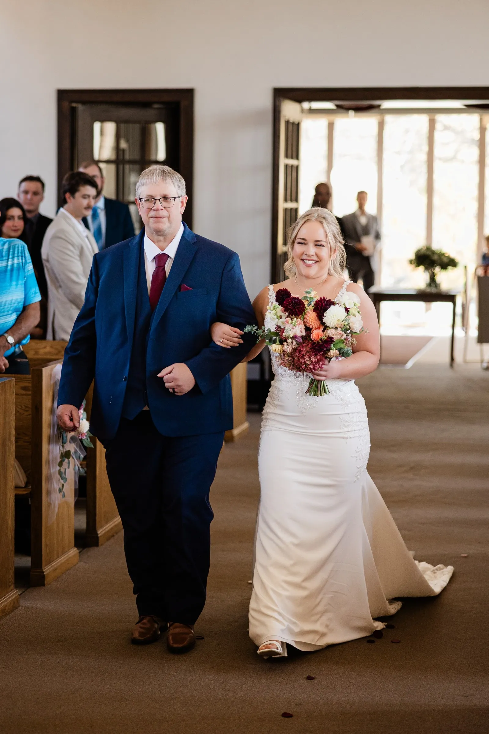 The bride smiles while walking arm in arm down the church aisle, carrying a burgundy and peach bouquet at St. Joseph Catholic Church in Crosby — Tim Larsen Photography, Brainerd Lakes MN