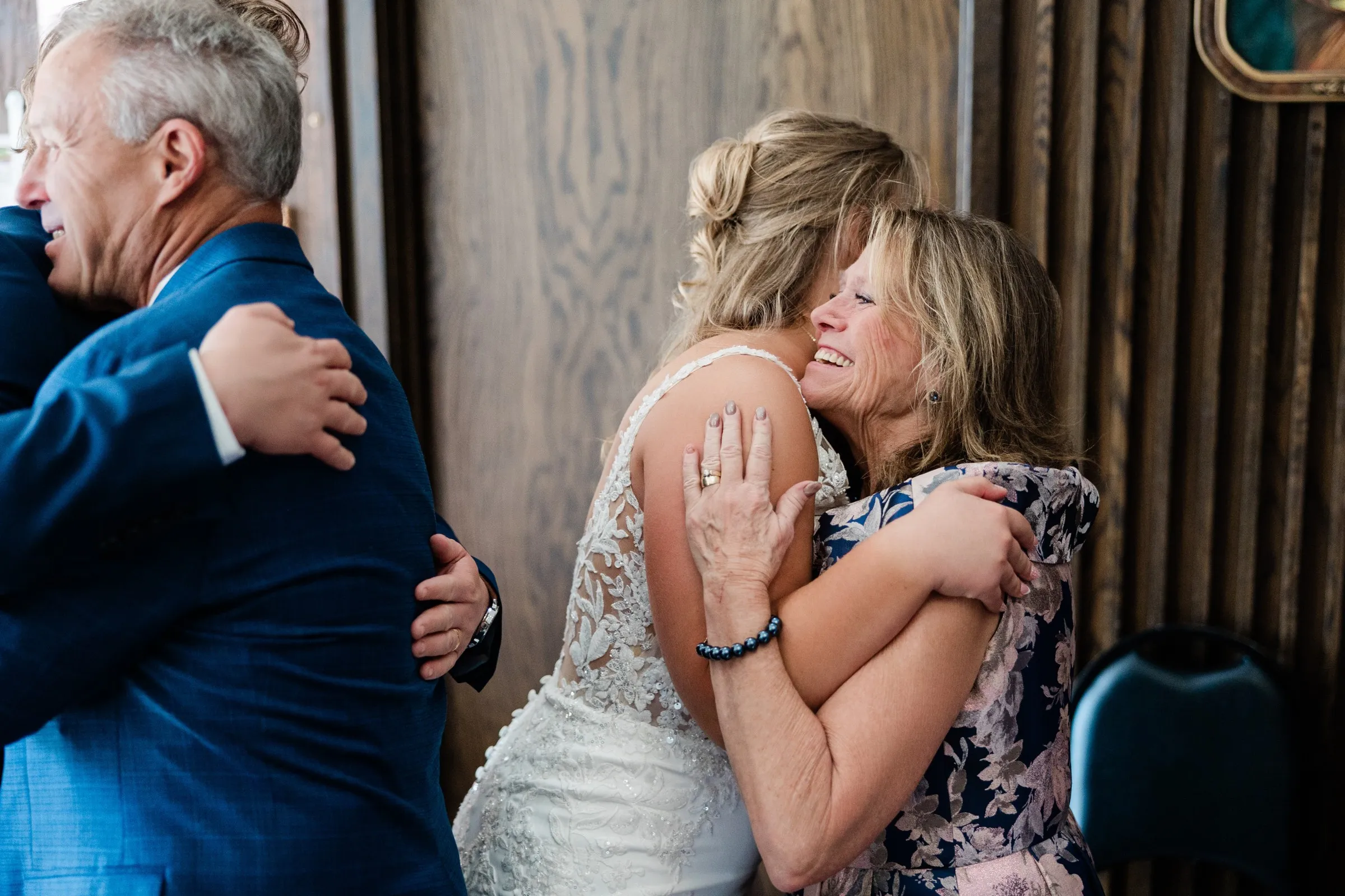 The bride embraces a smiling woman in a floral dress while the groom hugs another guest beside them at St. Joseph Catholic Church in Crosby — Tim Larsen Photography, Brainerd Lakes MN