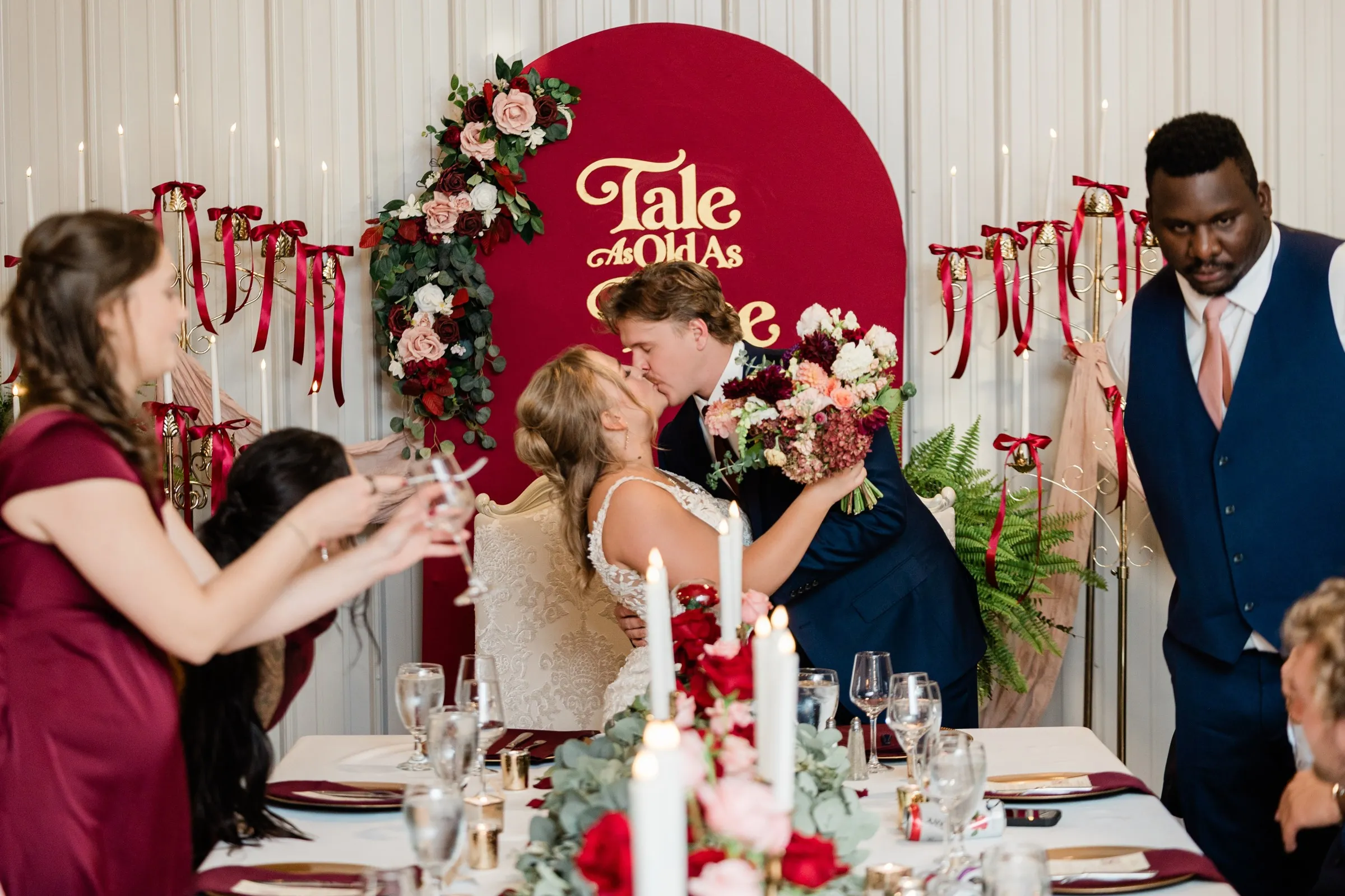 The couple kiss at the head table beneath a burgundy Tale As Old As Time sign decorated with roses at The Gathering in Crosslake — Tim Larsen Photography, Brainerd Lakes MN