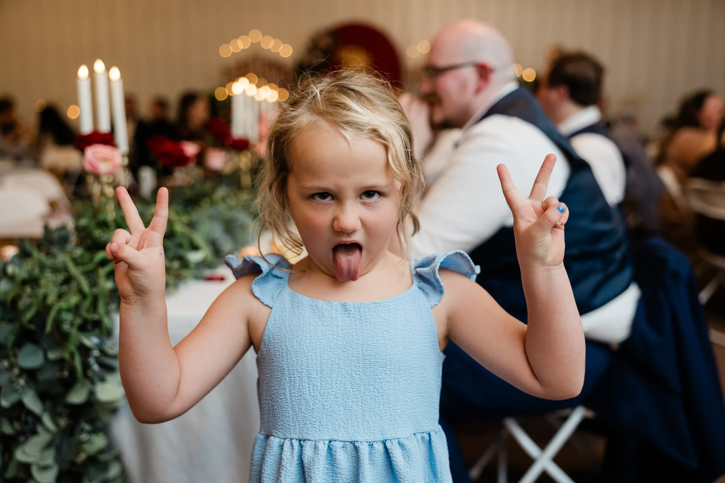A girl in a blue dress sticks out her tongue and raises two peace signs beside a reception table at The Gathering in Crosslake — Tim Larsen Photography, Brainerd Lakes MN