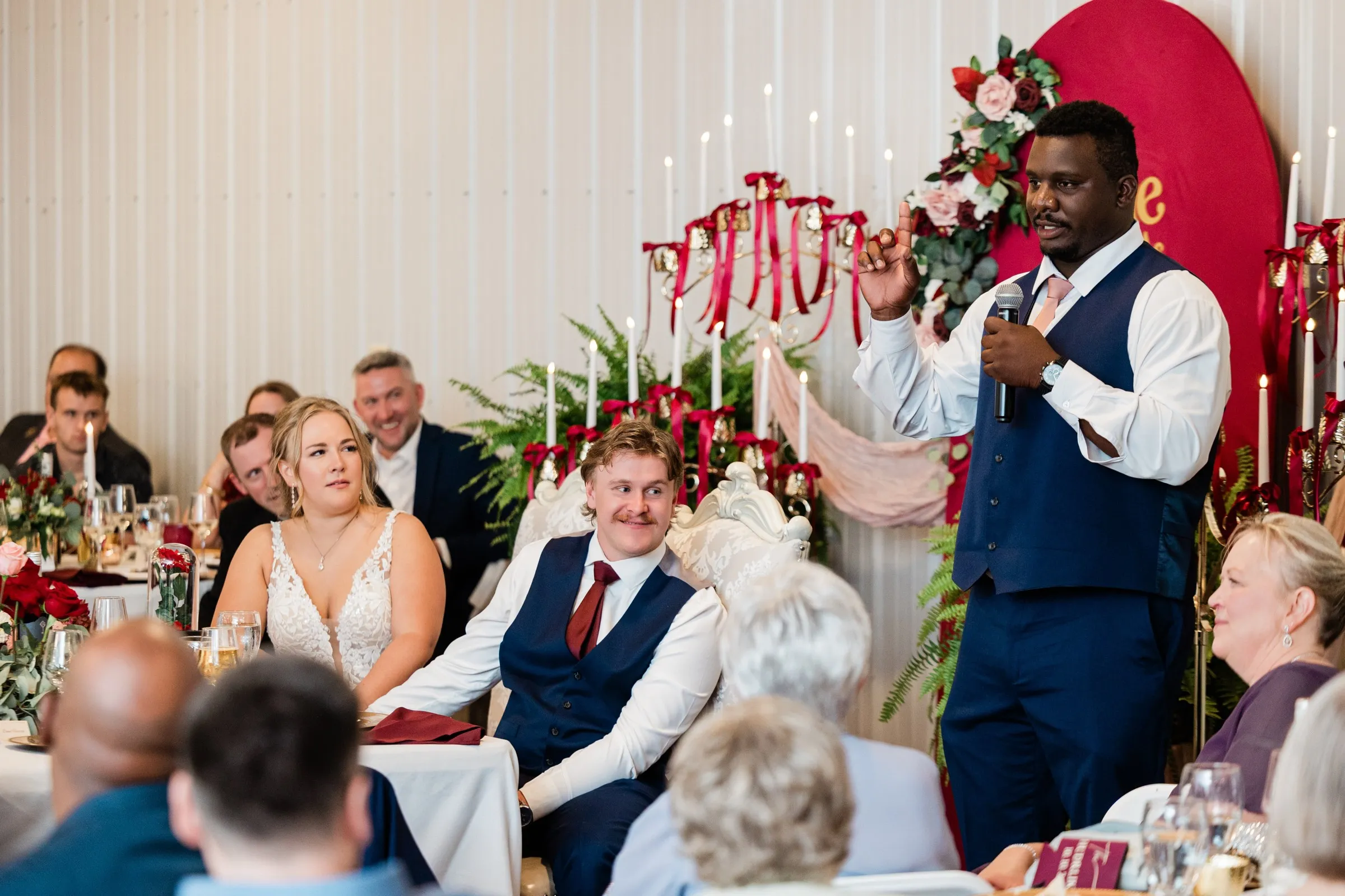 A wedding-party member speaks into a microphone beside the seated bride and groom at their candle-decorated head table at The Gathering in Crosslake — Tim Larsen Photography, Brainerd Lakes MN